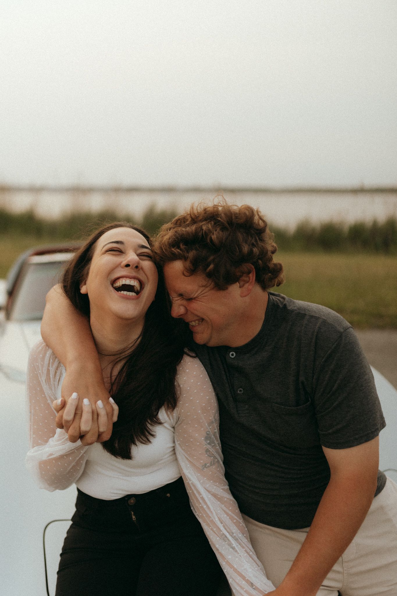 Laughing couple embracing while leaning on vintage car hood near open field and lake.