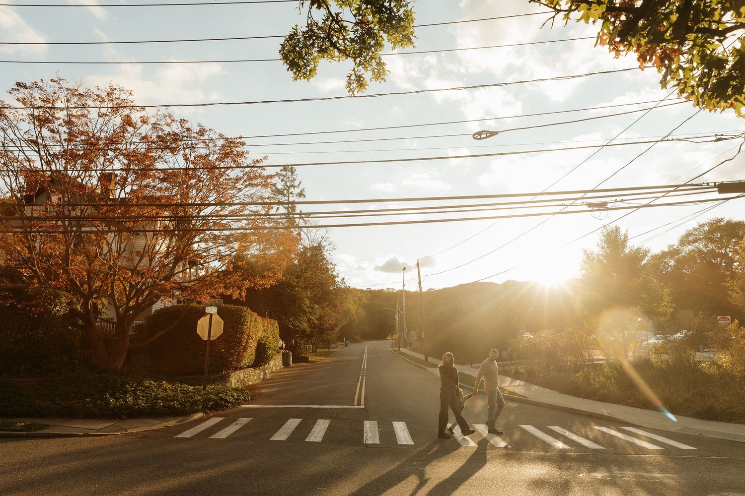People crossing a suburban street at sunset with power lines overhead.