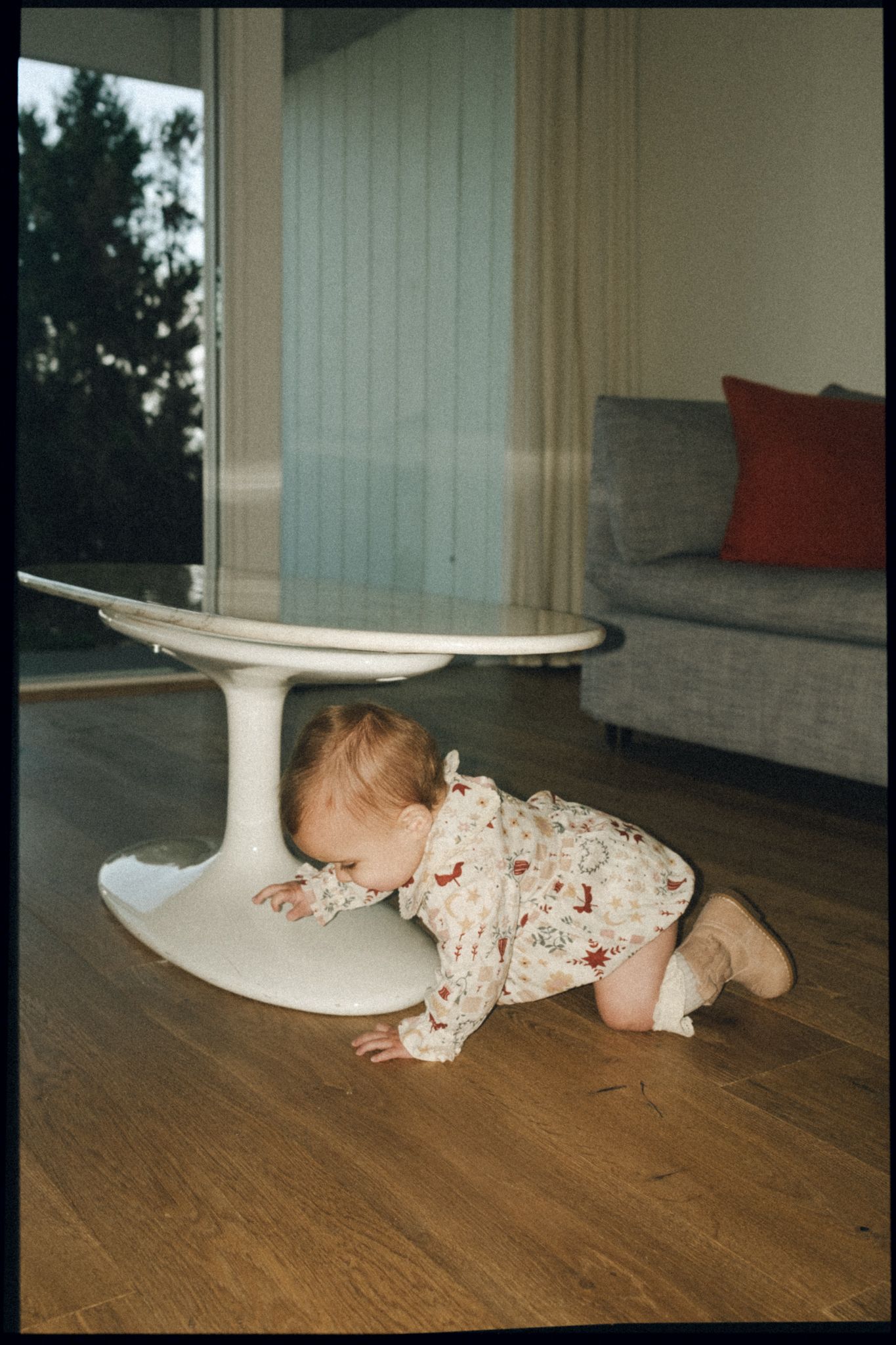 A curious baby in a floral outfit crawls under a white tulip table on hardwood floors in a bright living room.