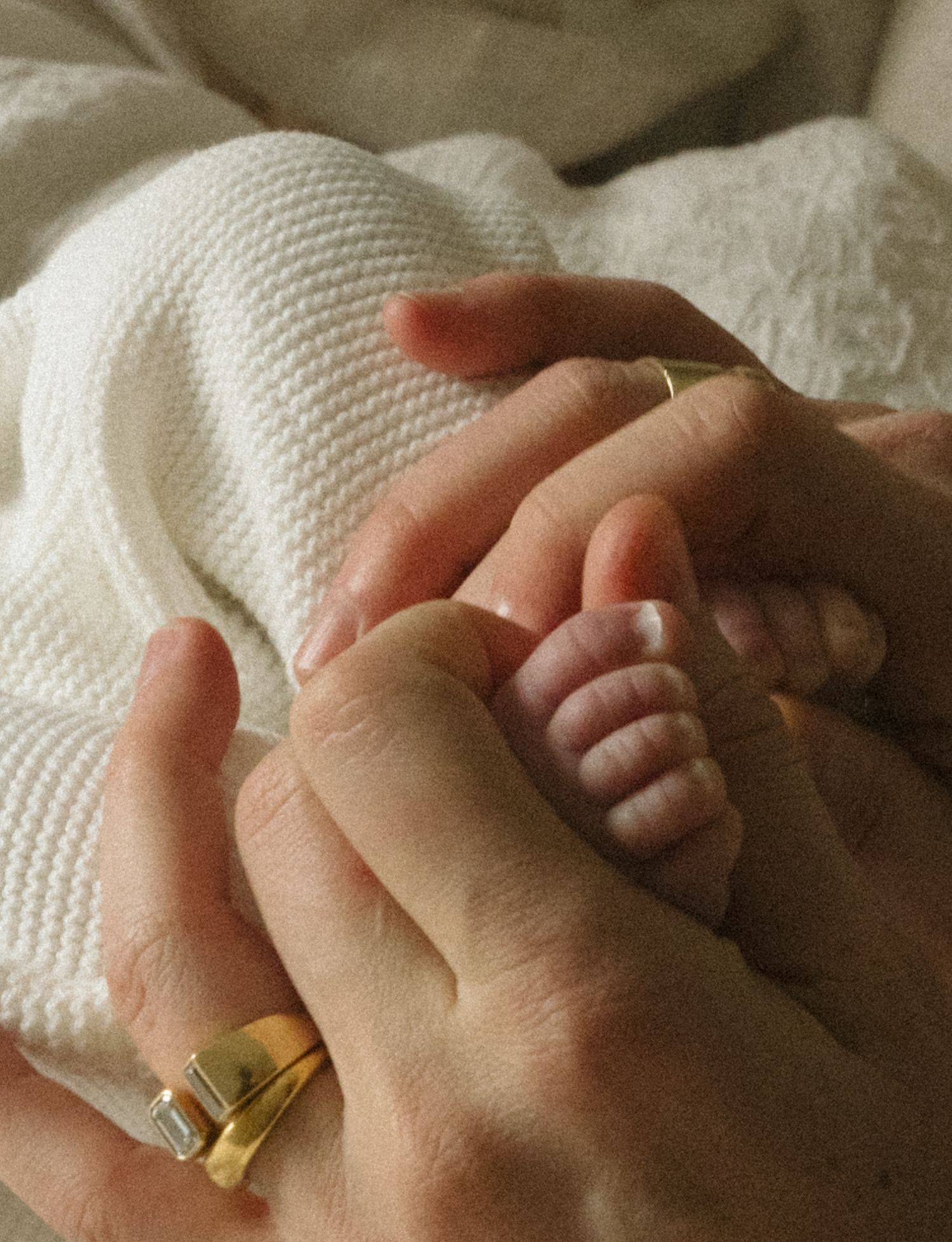 Adult hands gently holding a newborn baby's tiny feet wrapped in a white knit blanket, with a gold ring visible.