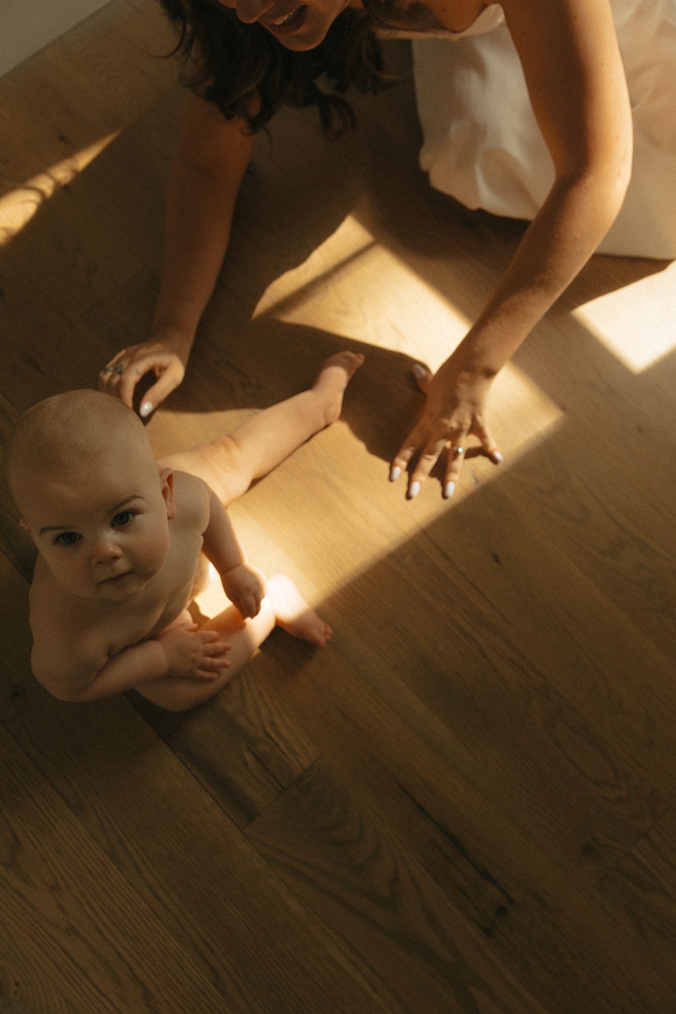 A baby sits on a warm wooden floor in golden light while a woman reaches toward them from behind.