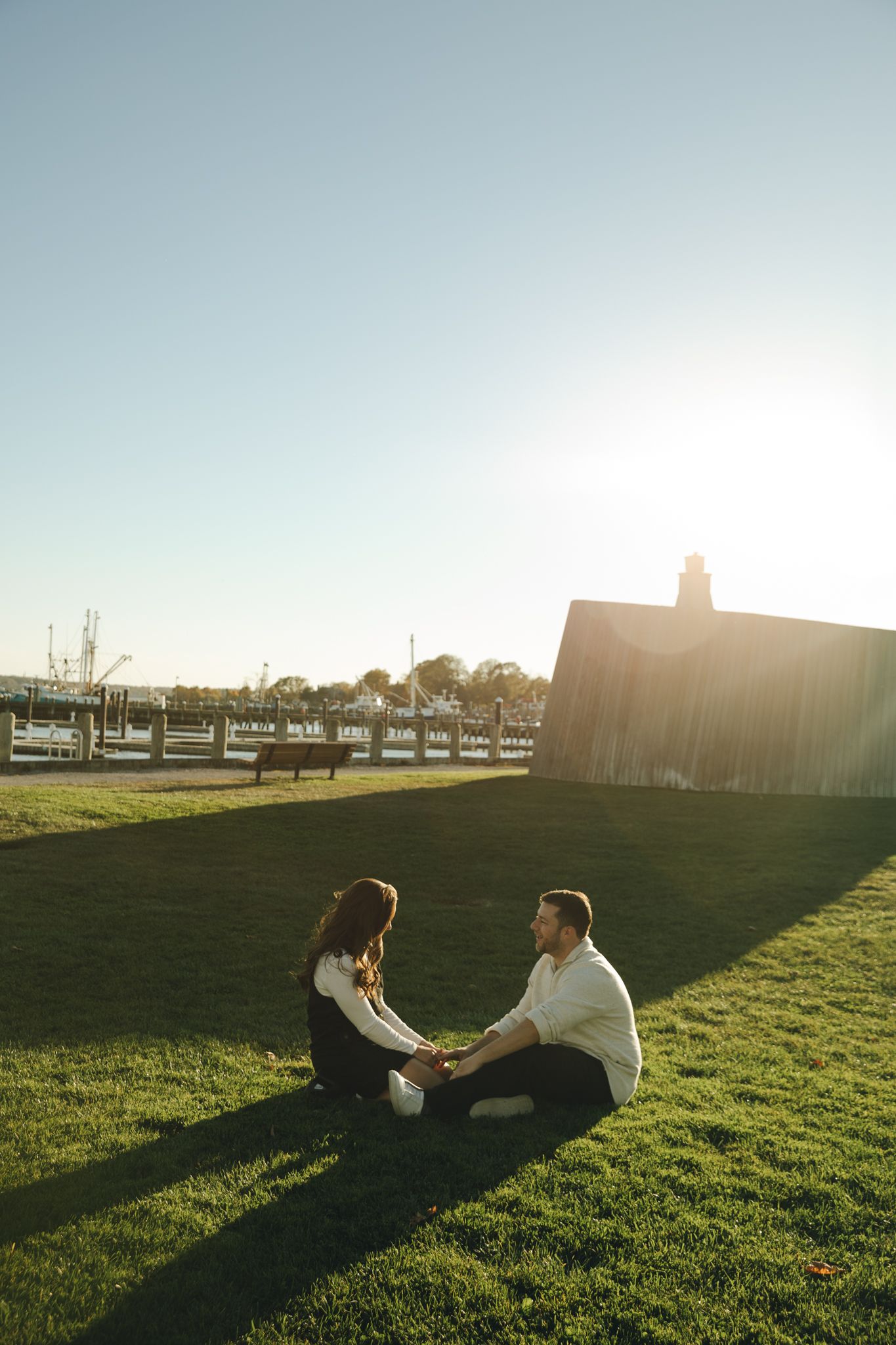 Couple sitting on green grass holding hands at golden hour with marina and small building in background.