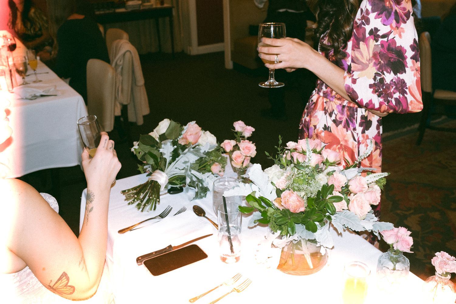 Elegant wedding reception table with pink floral centerpieces, gold cutlery, and guests in floral dress holding wine glass.