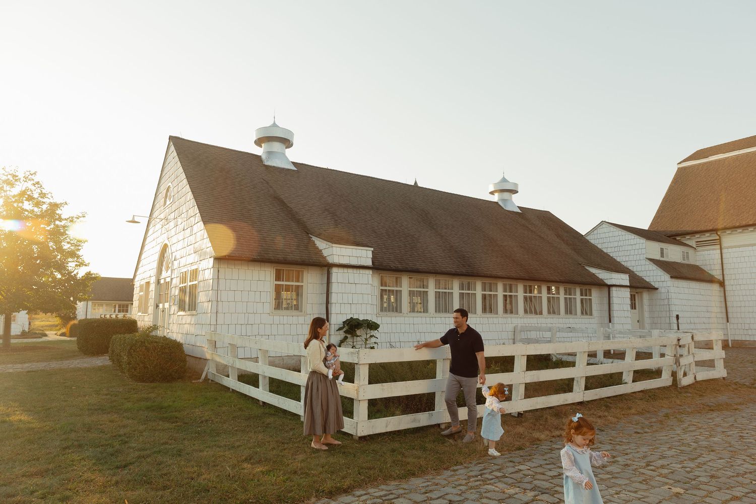 Family standing outside a charming white barn building with white fence at golden hour sunset.