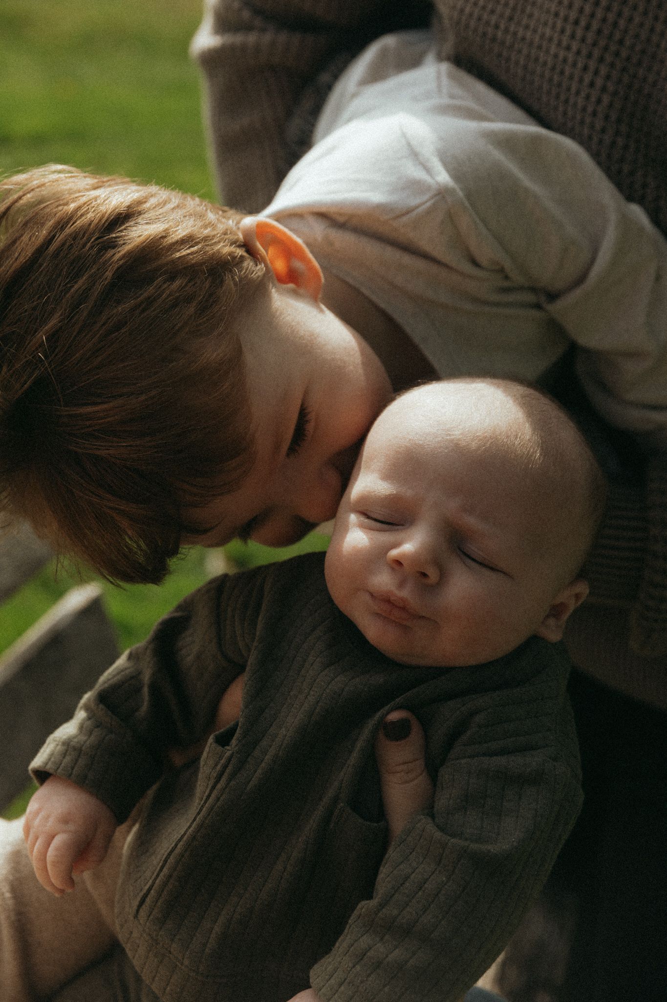 Young child gently kissing a sleeping newborn baby sibling held by an adult outdoors in natural light.