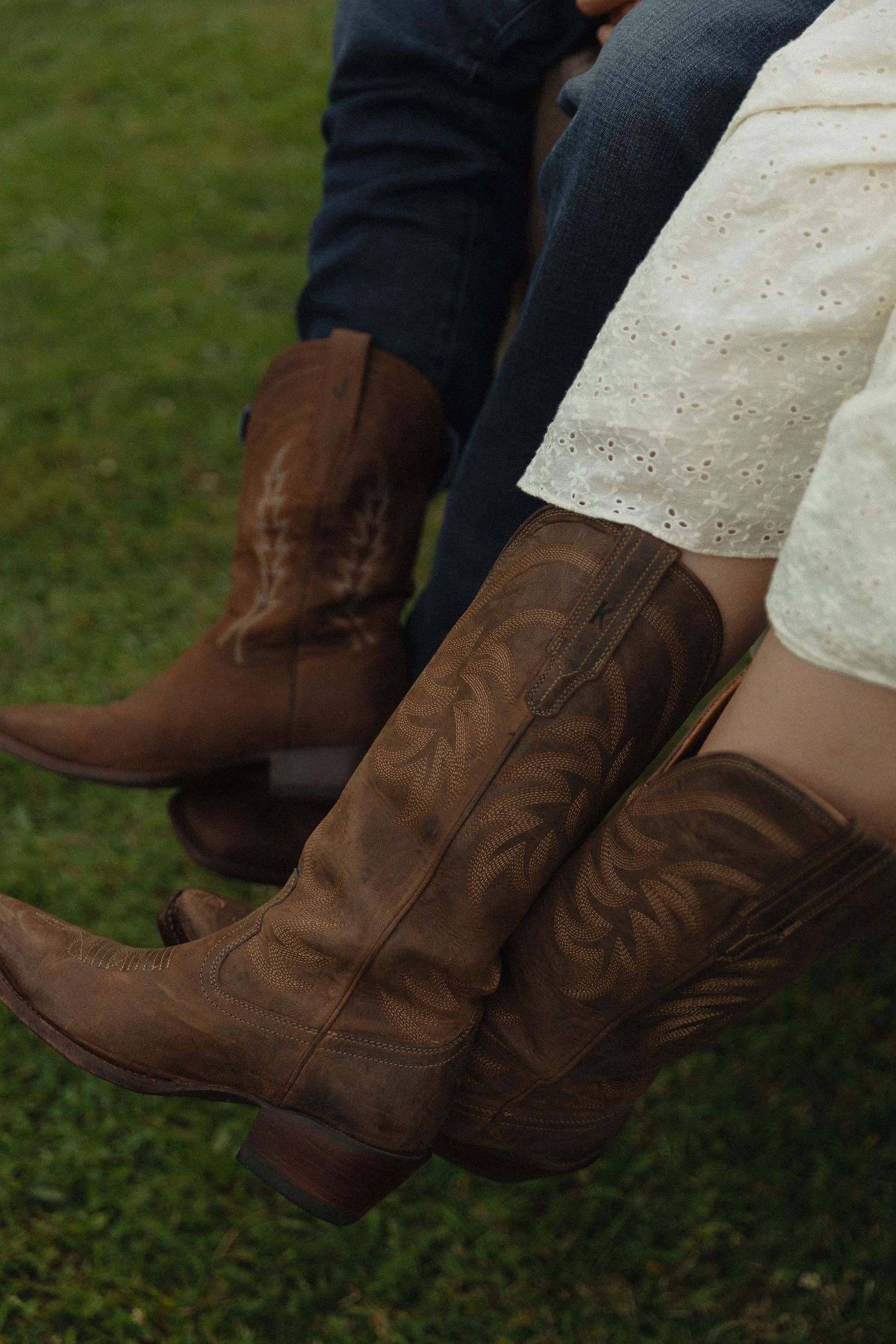 Two people wearing matching brown leather cowboy boots sitting on green grass, one in jeans and one in a white lace skirt.