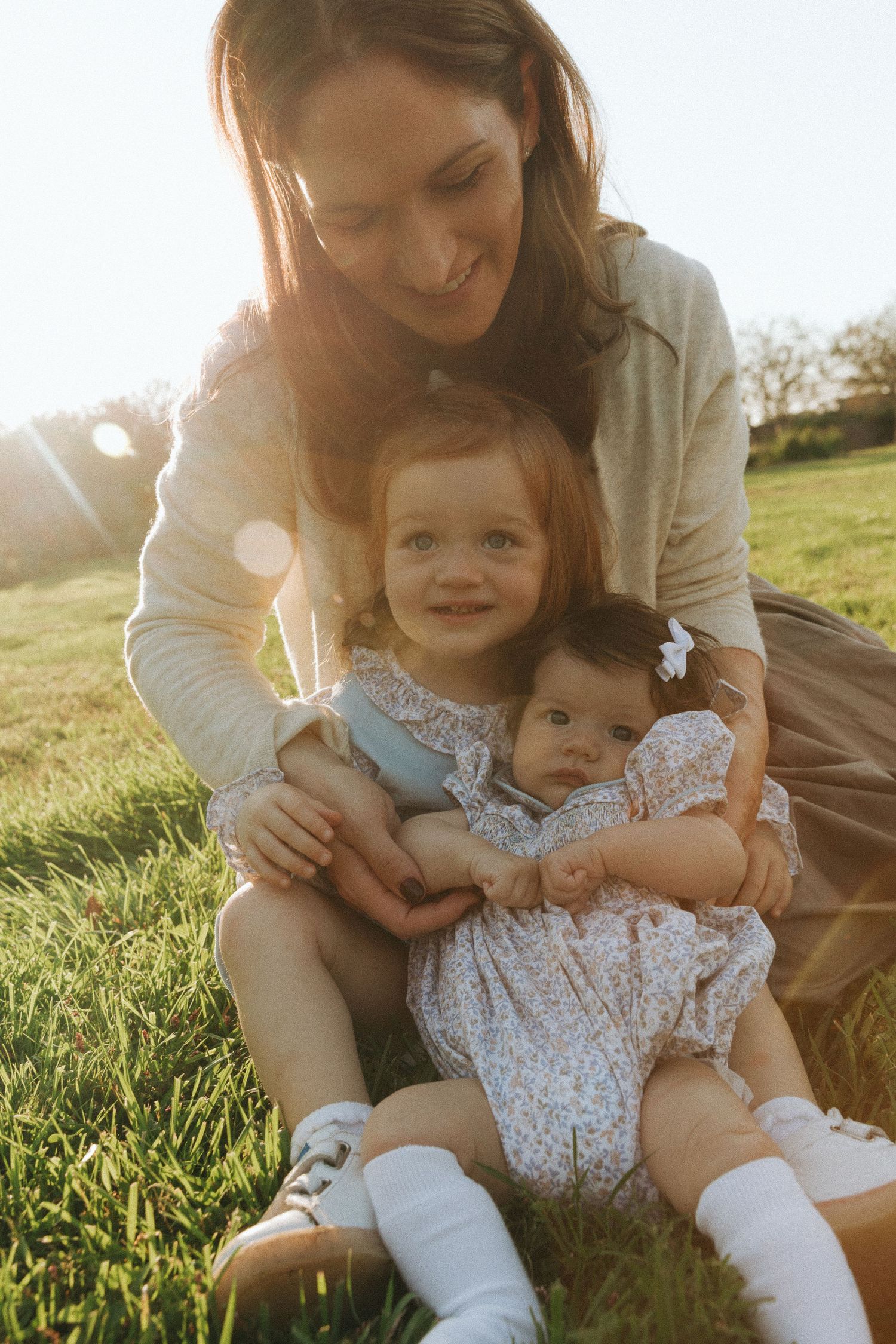A mother sits on green grass holding two young daughters in golden sunlight outdoors.