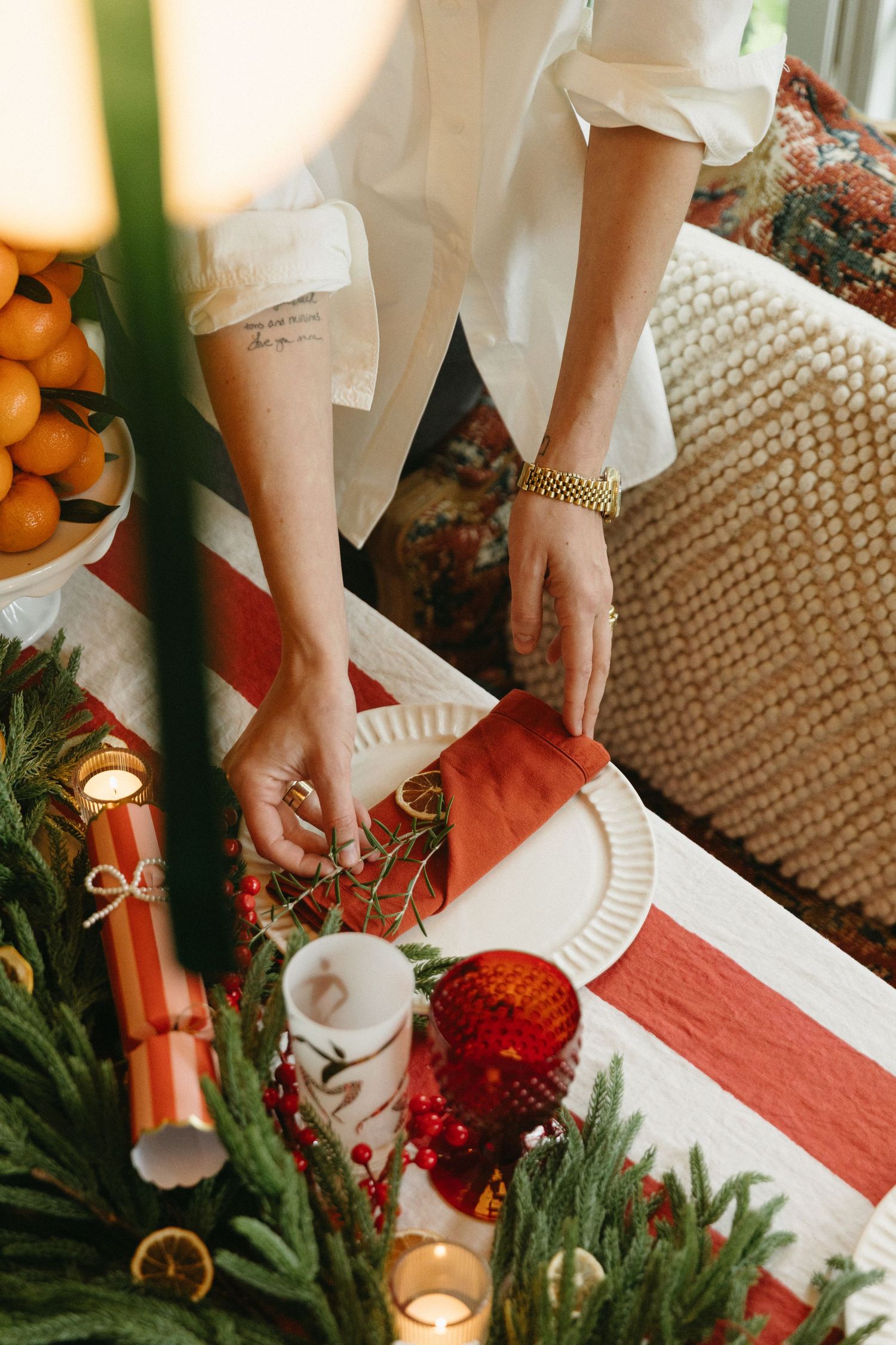 Person arranging festive Christmas table setting with red napkins, greenery, candles, and holiday decorations.