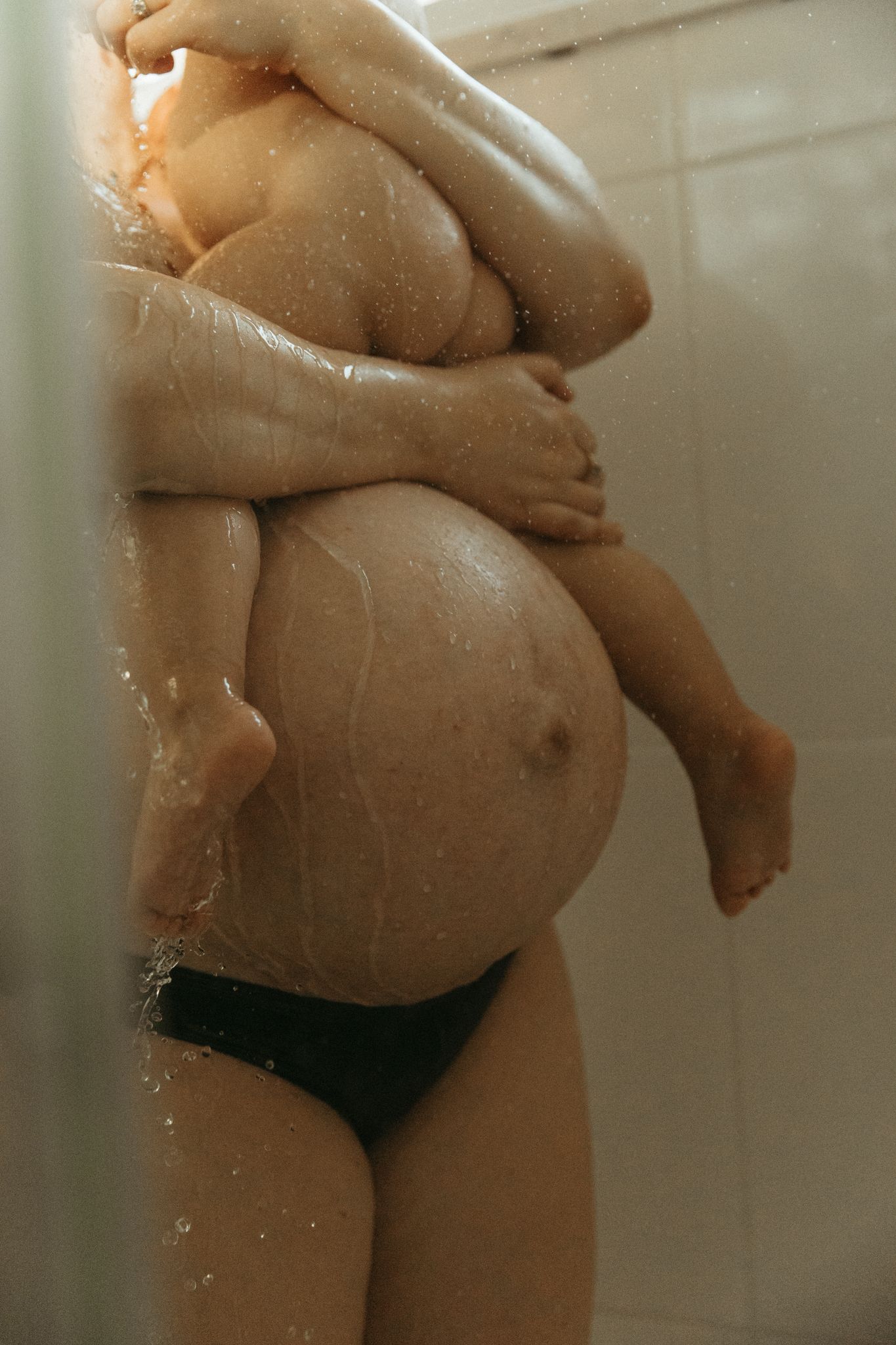 Pregnant woman in black underwear embracing her baby bump while showering in a steamy bathroom.