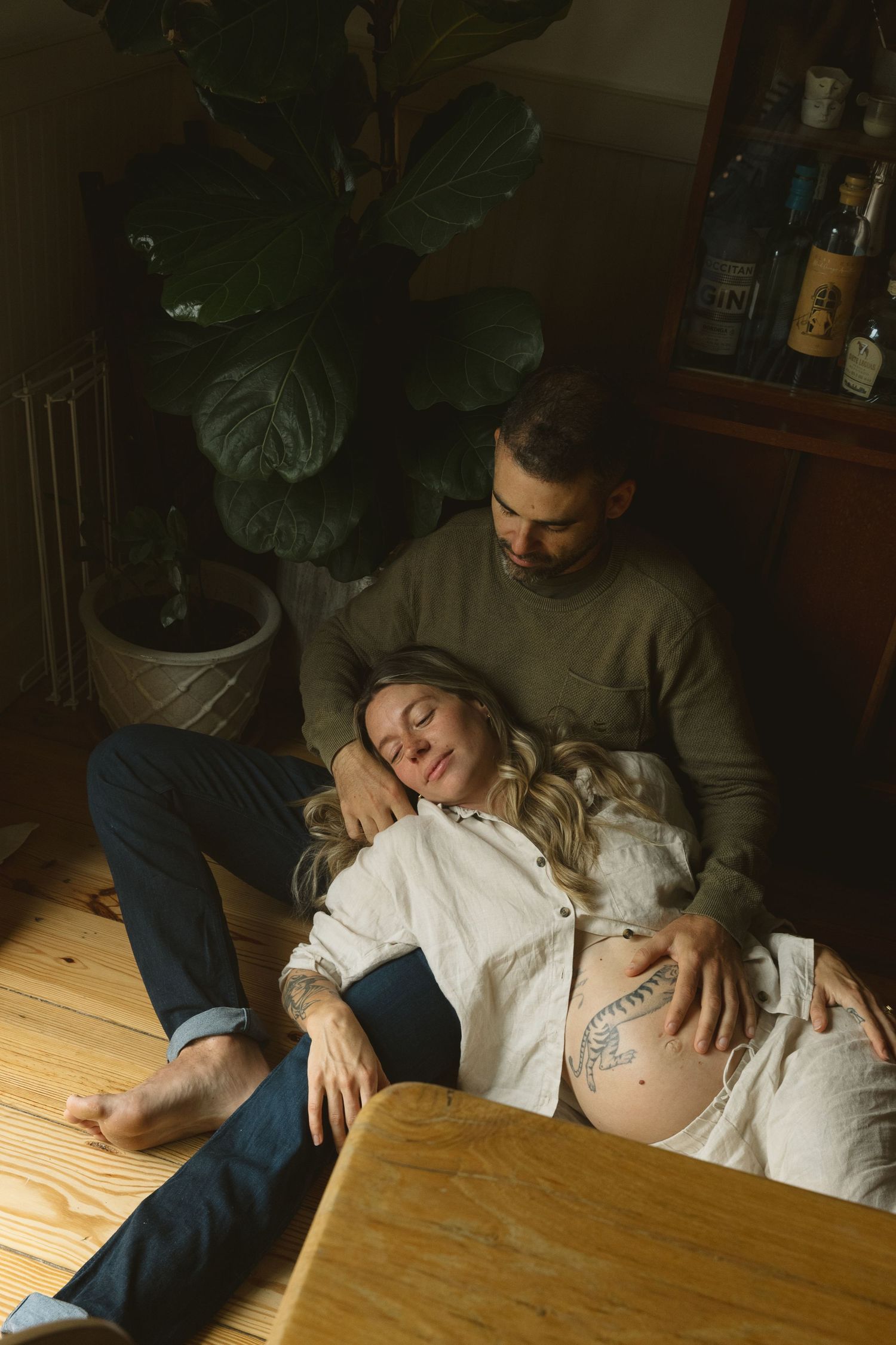 Couple relaxing on floor, pregnant woman leaning on partner near wooden table and large plant indoors.