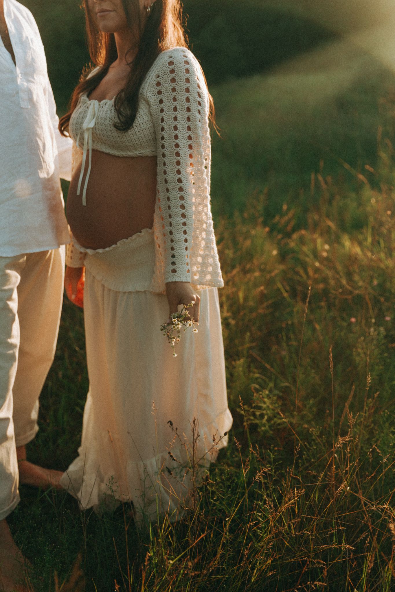 Pregnant woman in white crochet top and skirt holds wildflowers in sunlit green meadow.