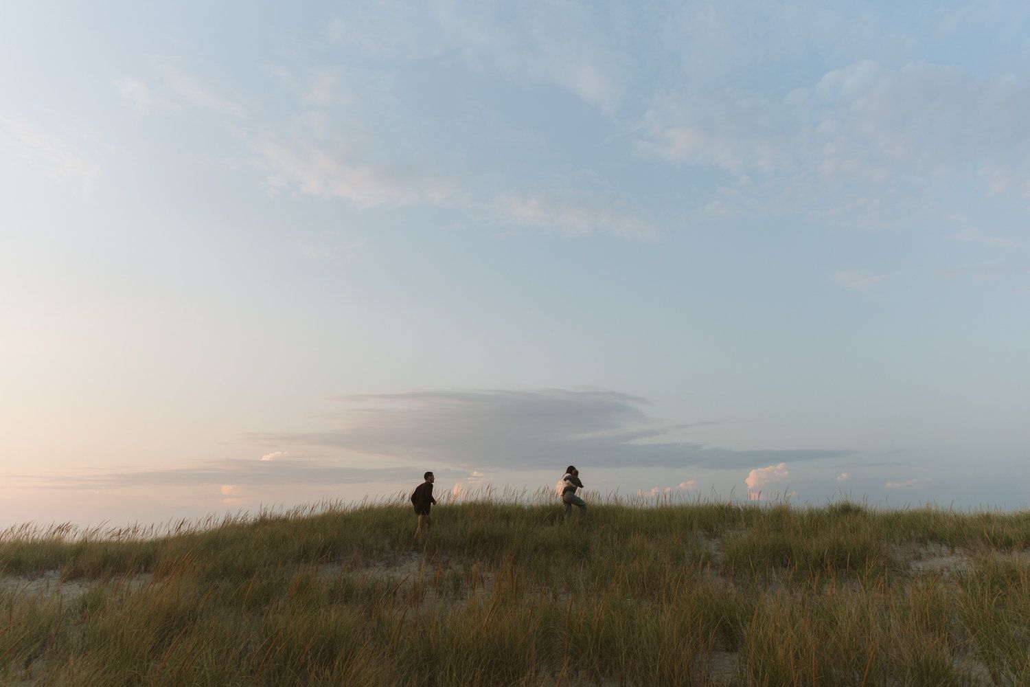 Two people walking on grassy sand dunes under a pastel evening sky.