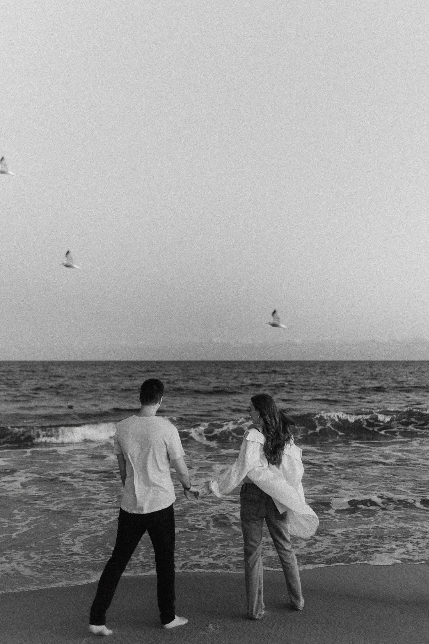 A couple enjoys a playful moment on a beach with seagulls flying overhead in black and white.