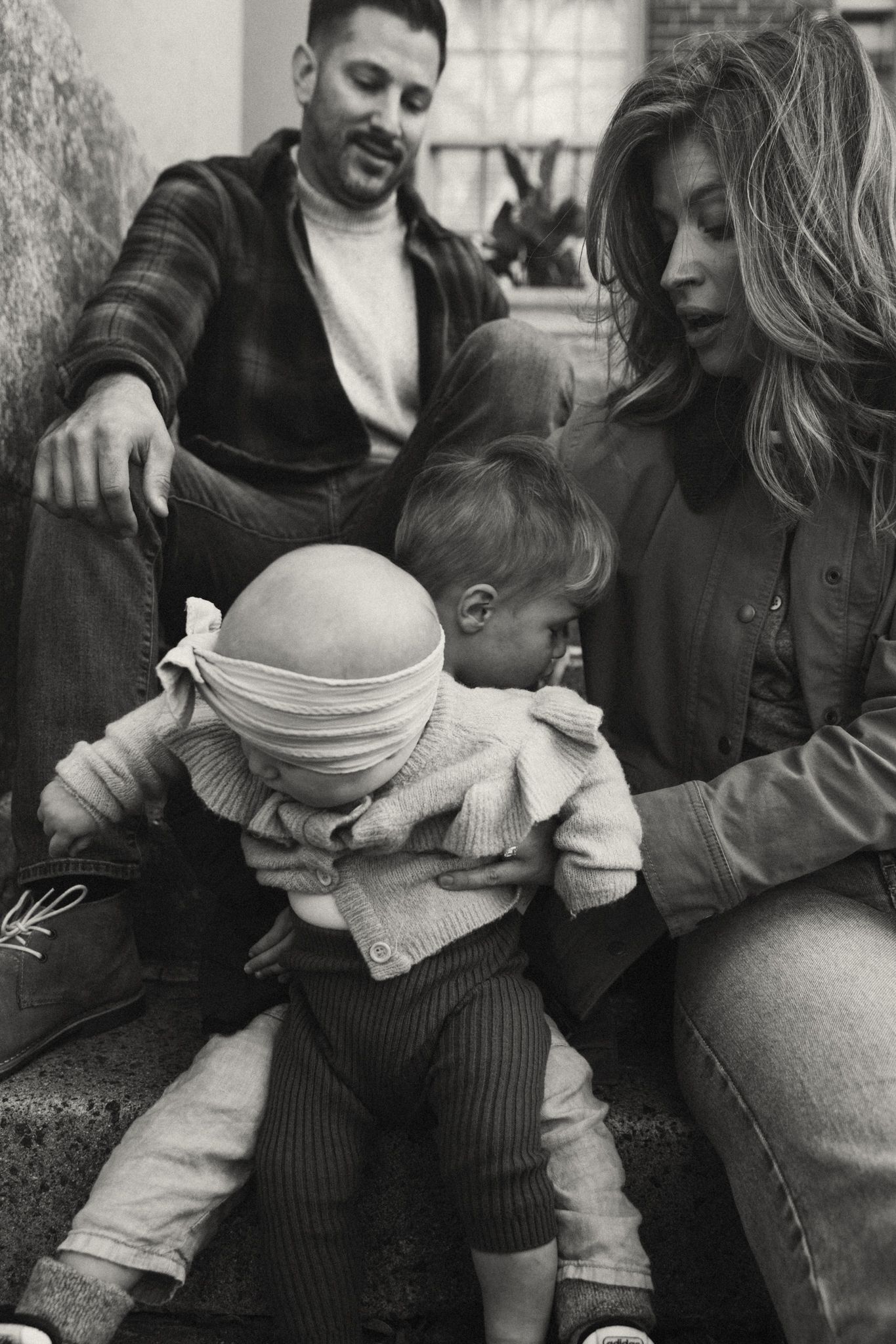 Black and white family photo of parents sitting with two young children on steps in a cozy indoor setting.