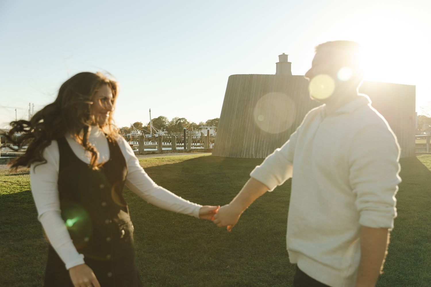 Couple holding hands outdoors in golden sunset light, woman in black dress, man in white sweater on green grass.