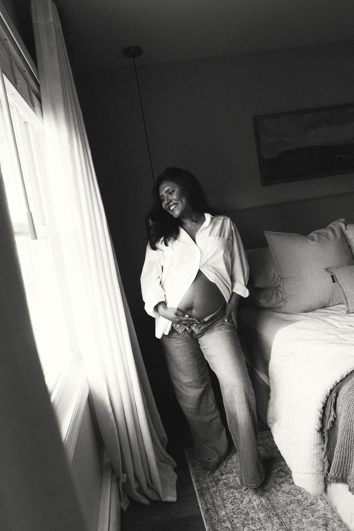 Black and white photo of a smiling pregnant woman in jeans and open white shirt standing by a sunlit bedroom window.