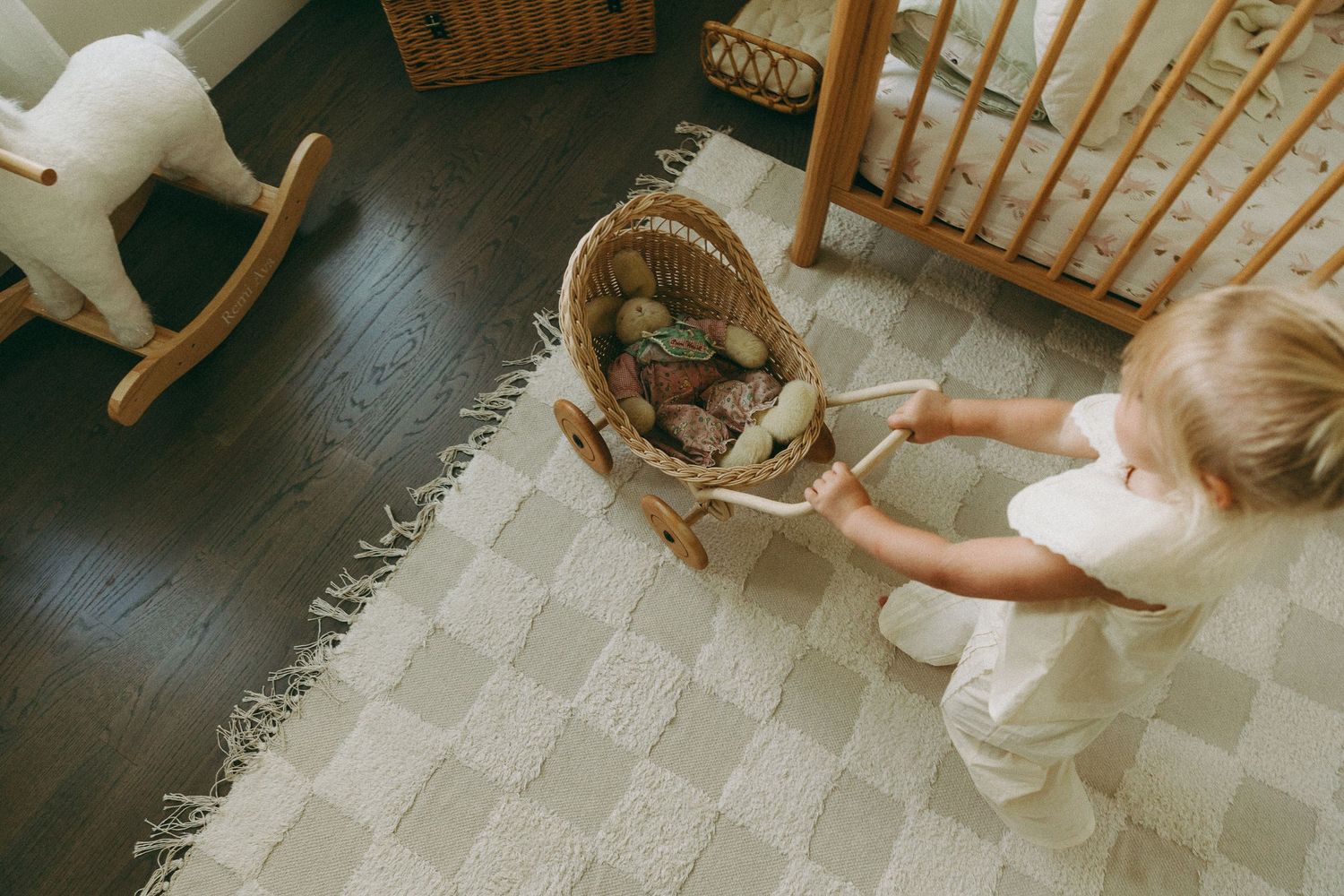 Toddler girl pushing a wicker doll pram in a cozy nursery with a wooden crib and rocking horse on a checkered rug.