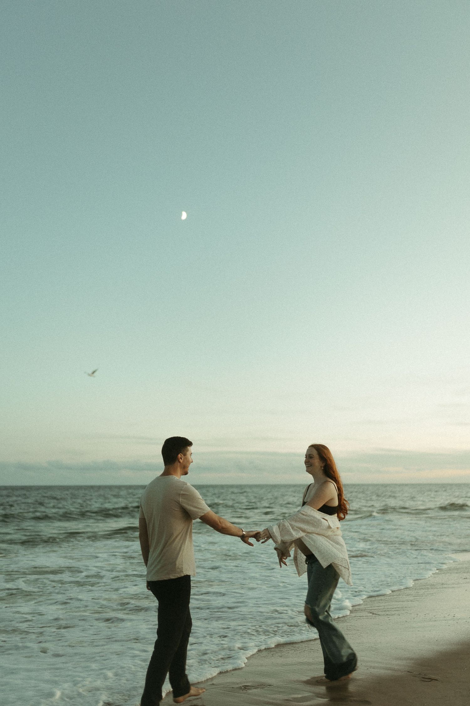 Two people hold hands and dance together on a serene beach at sunset.
