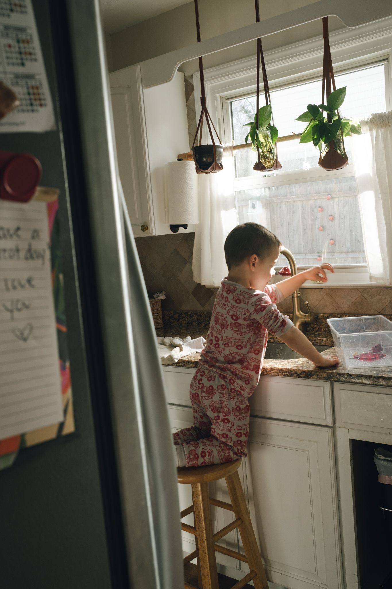 A child in patterned pajamas stands on a wooden stool at a kitchen sink with hanging plants in the window above.
