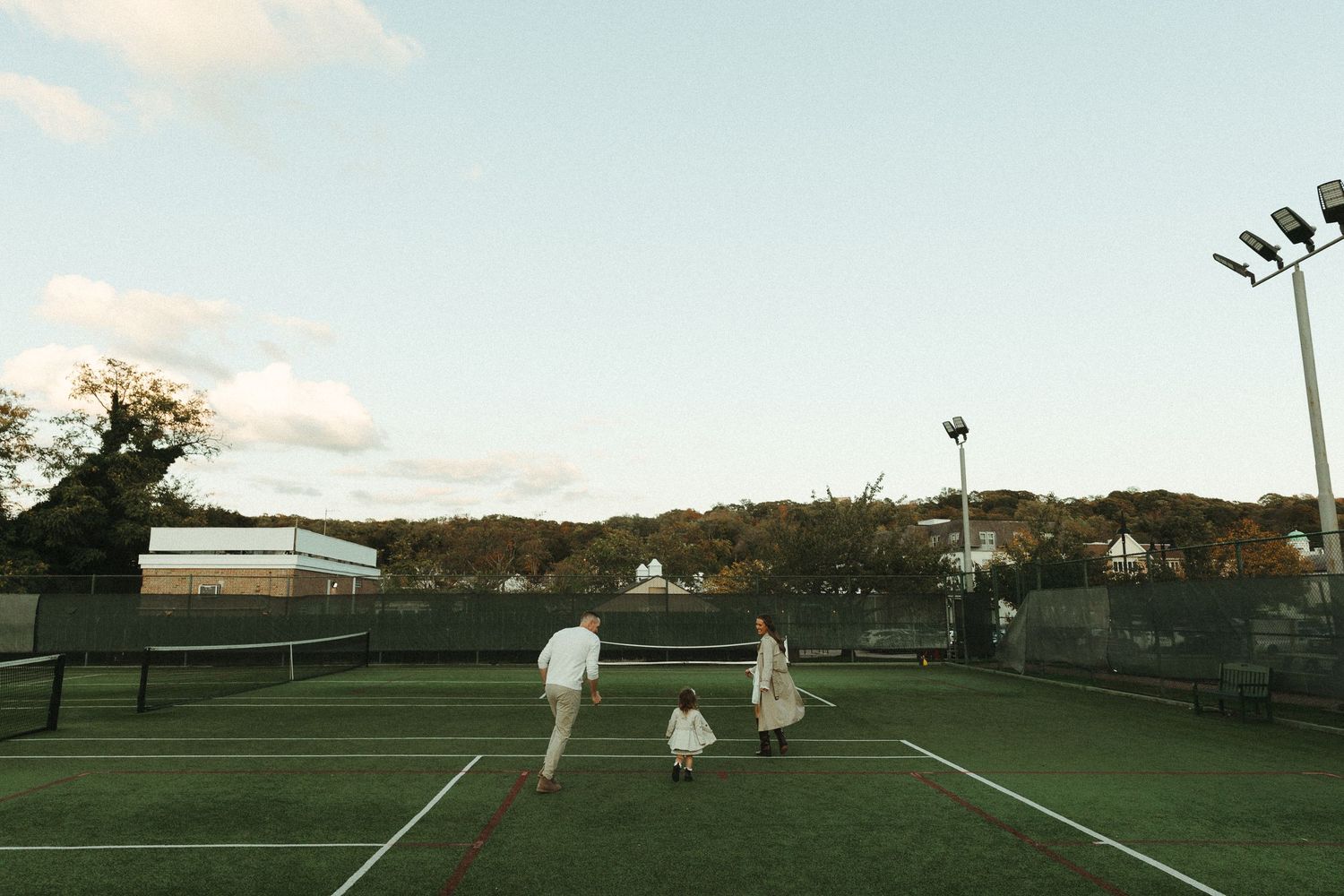 A tennis court surrounded by trees at dusk with floodlights illuminating the playing surface.