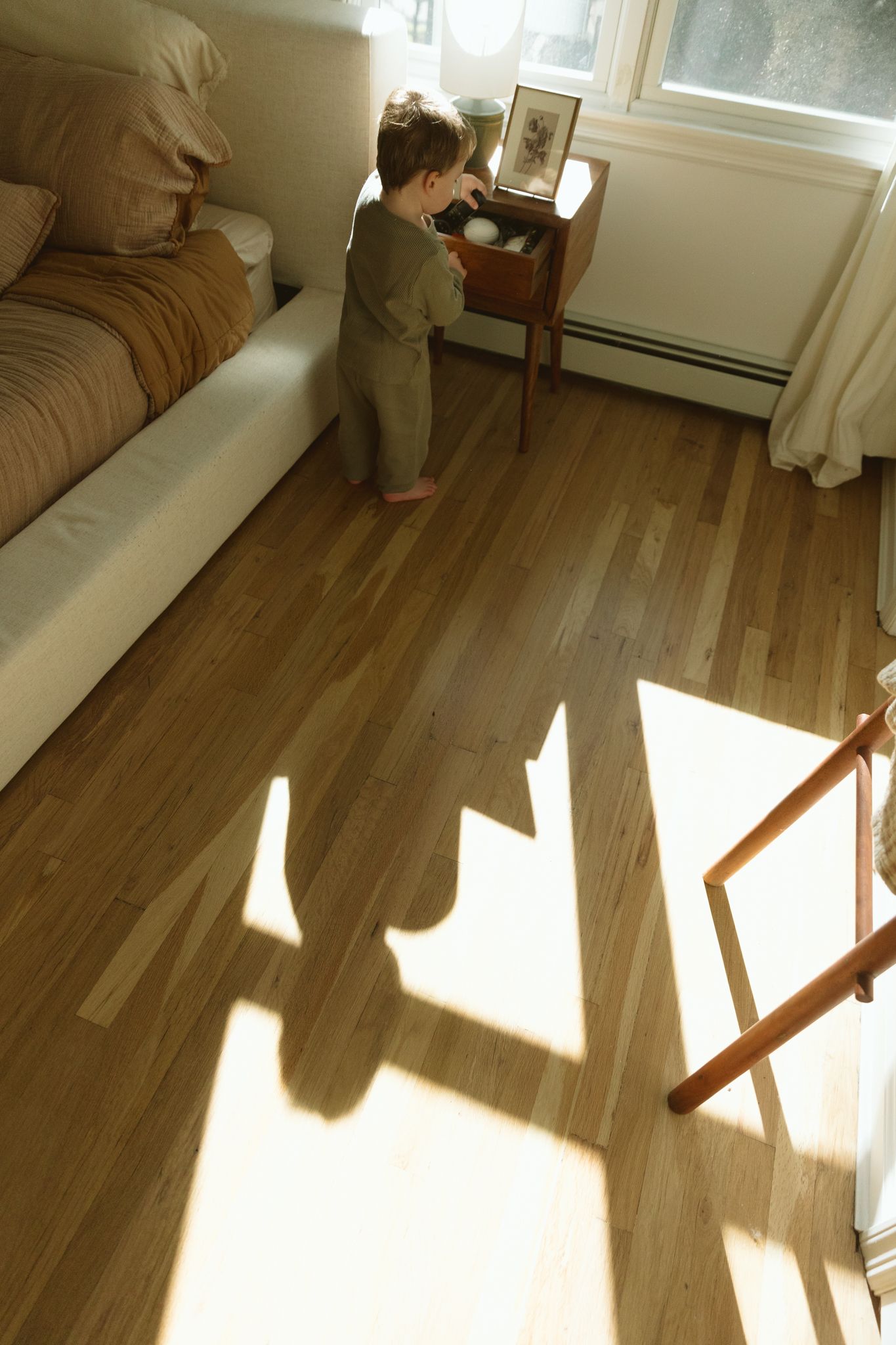 A toddler in green pajamas stands on sunny hardwood floors beside a bedside table, casting a long shadow in a cozy bedroom.