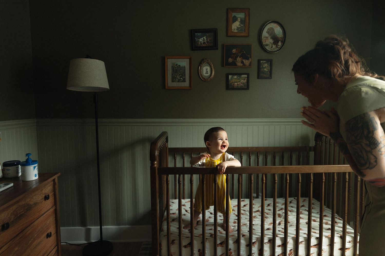 Smiling baby in wooden crib with tattooed parent leaning over in cozy nursery with framed photos on green wall.