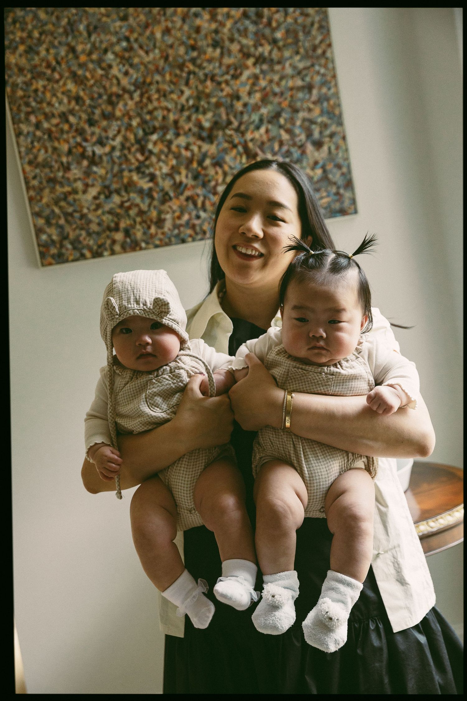 Smiling Asian woman holding two adorable babies dressed in matching outfits with white socks indoors.