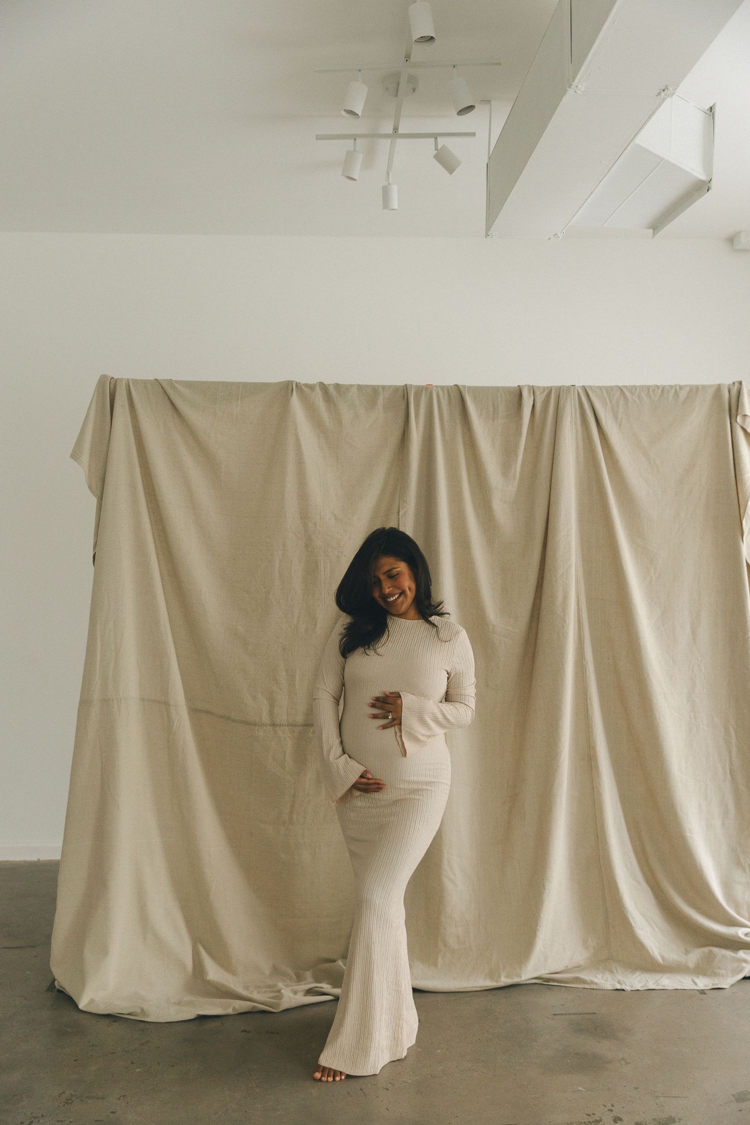 Pregnant woman in white fitted maxi dress smiling and posing in front of beige fabric backdrop in studio.