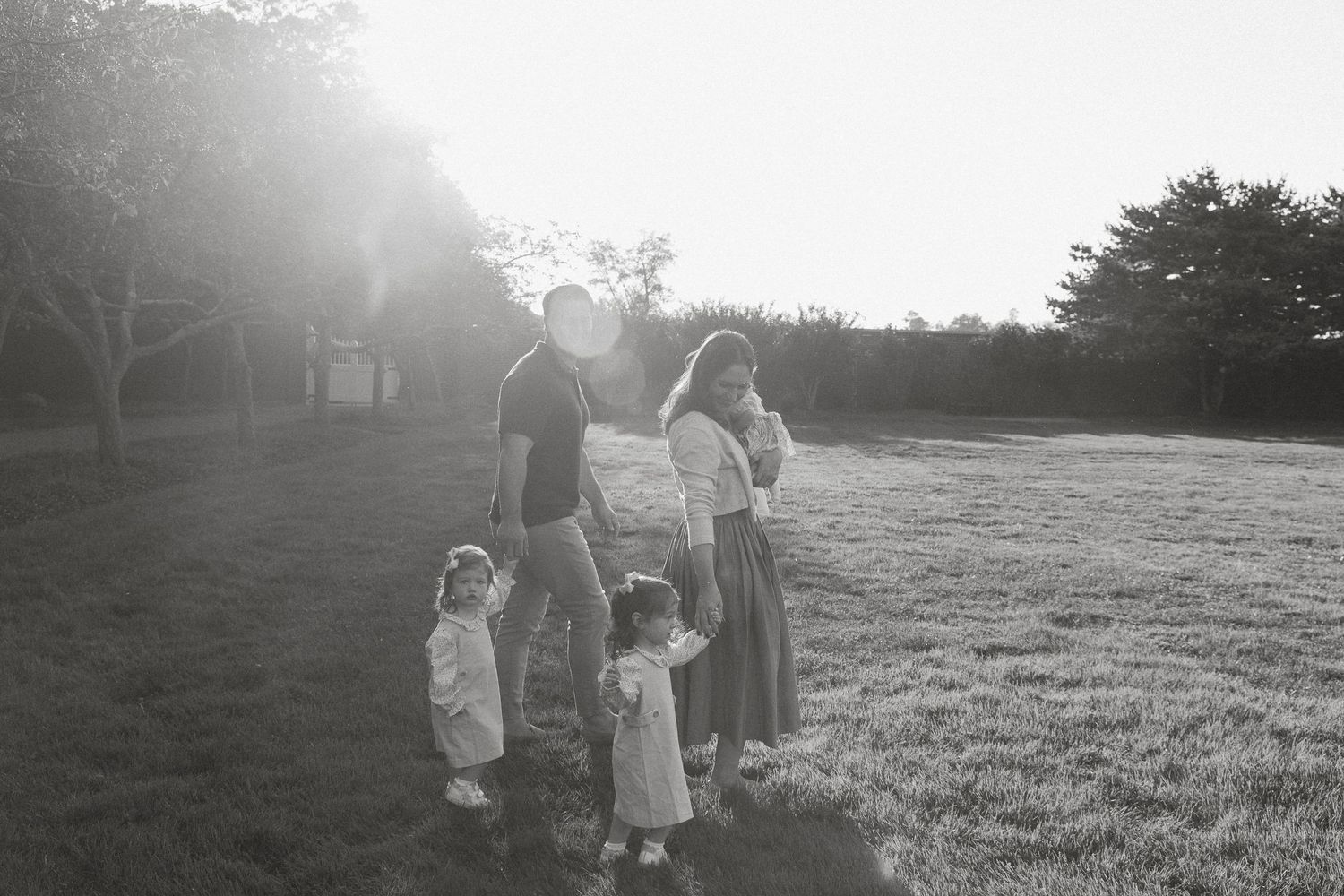 Black and white family photo of parents with two young children standing in a sunlit grassy field at golden hour.