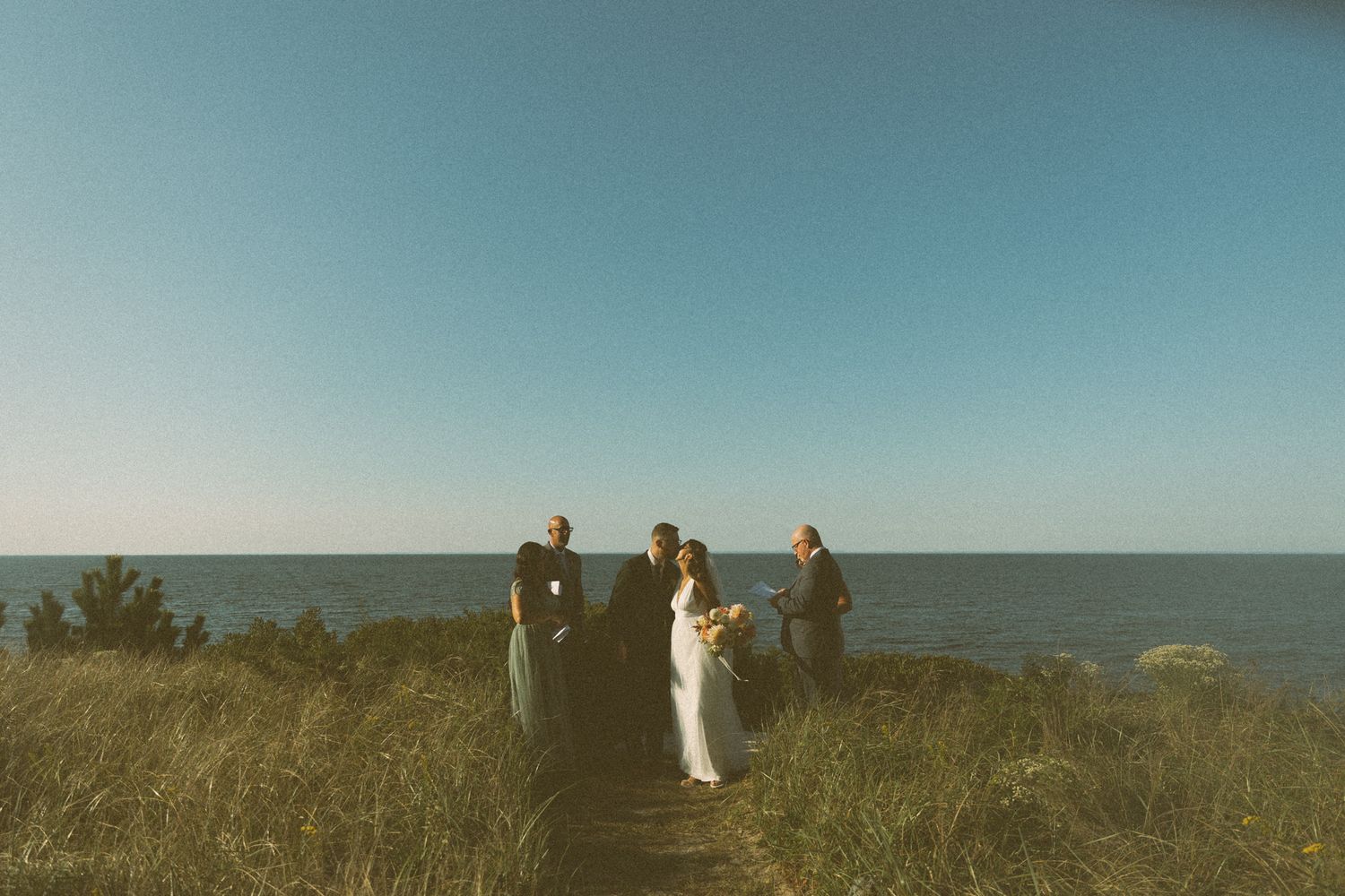 Outdoor wedding ceremony on a wooden boardwalk overlooking the ocean, with couple and officiant surrounded by golden grass.