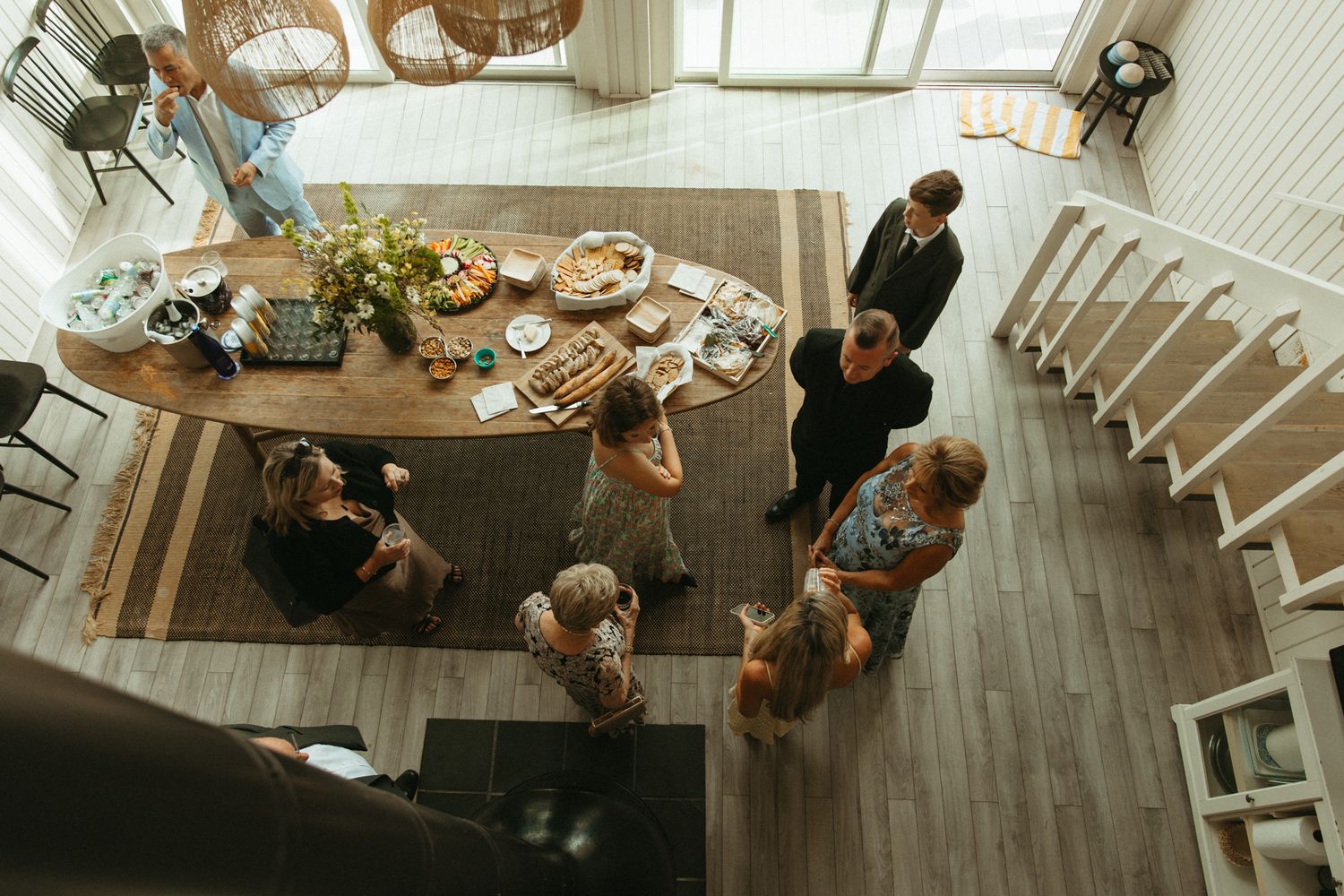 Aerial view of a casual indoor gathering with people socializing around a wooden dining table with food and drinks.