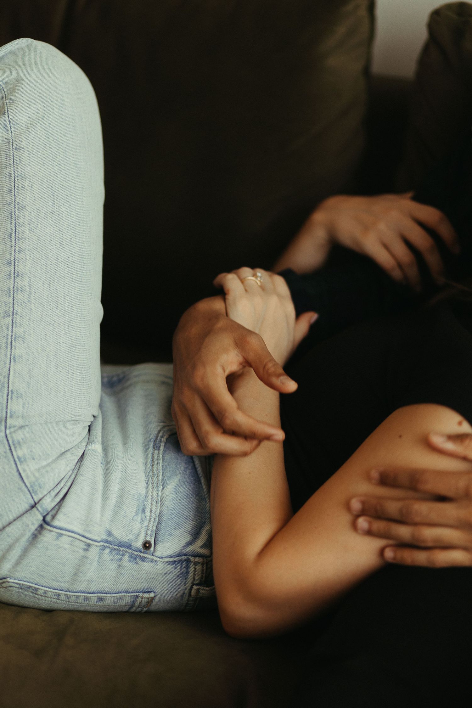 Close-up of couple holding hands on dark couch, woman wearing engagement ring in jeans.