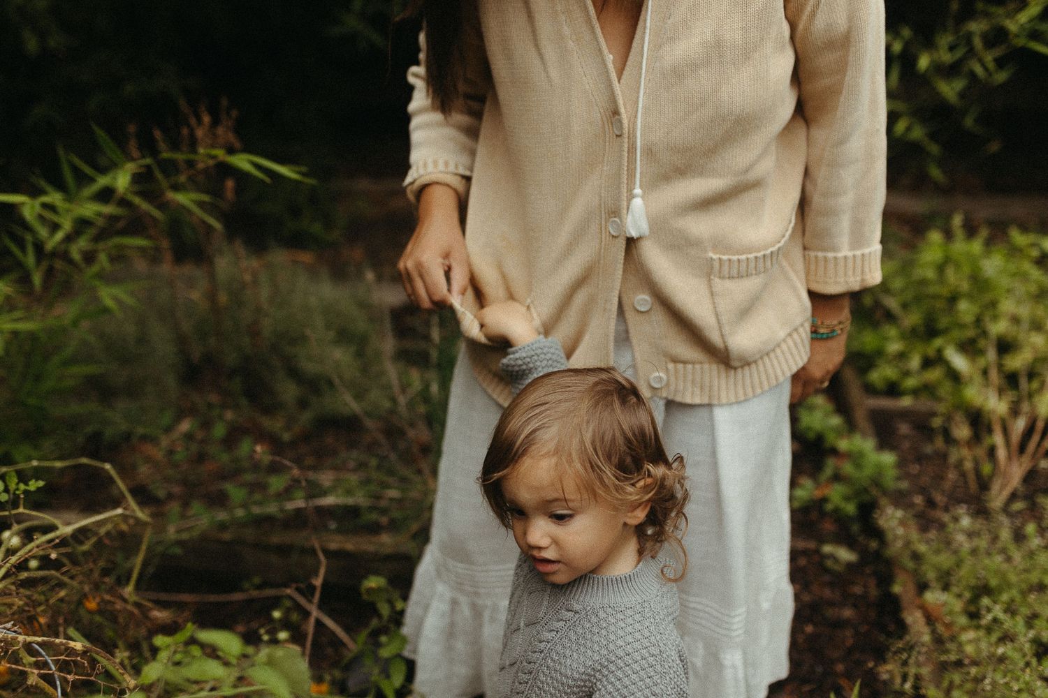 Young girl in gray knit sweater holding mother's hand in a lush garden setting.