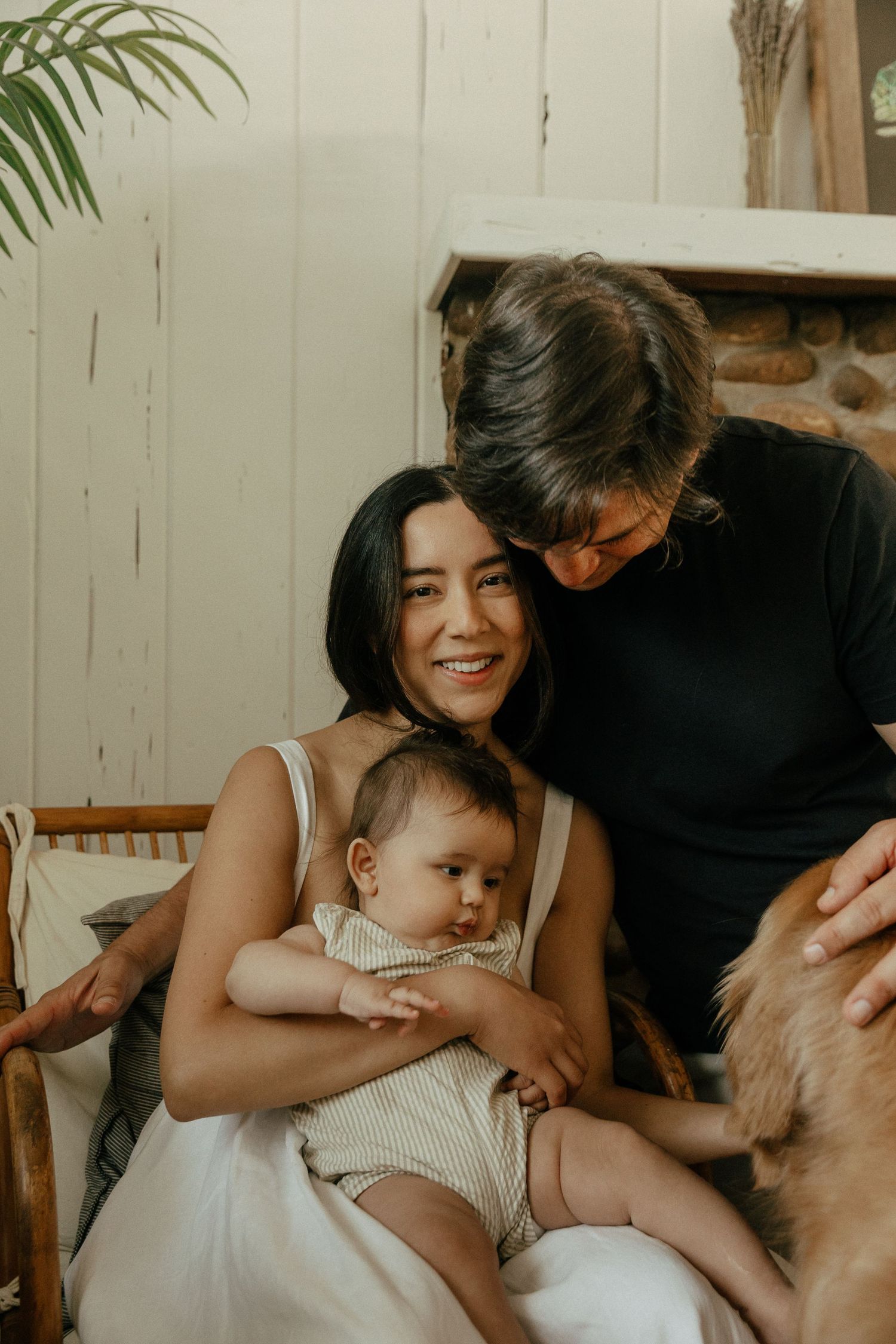 Smiling mother holding newborn baby with partner leaning in beside a stone fireplace.