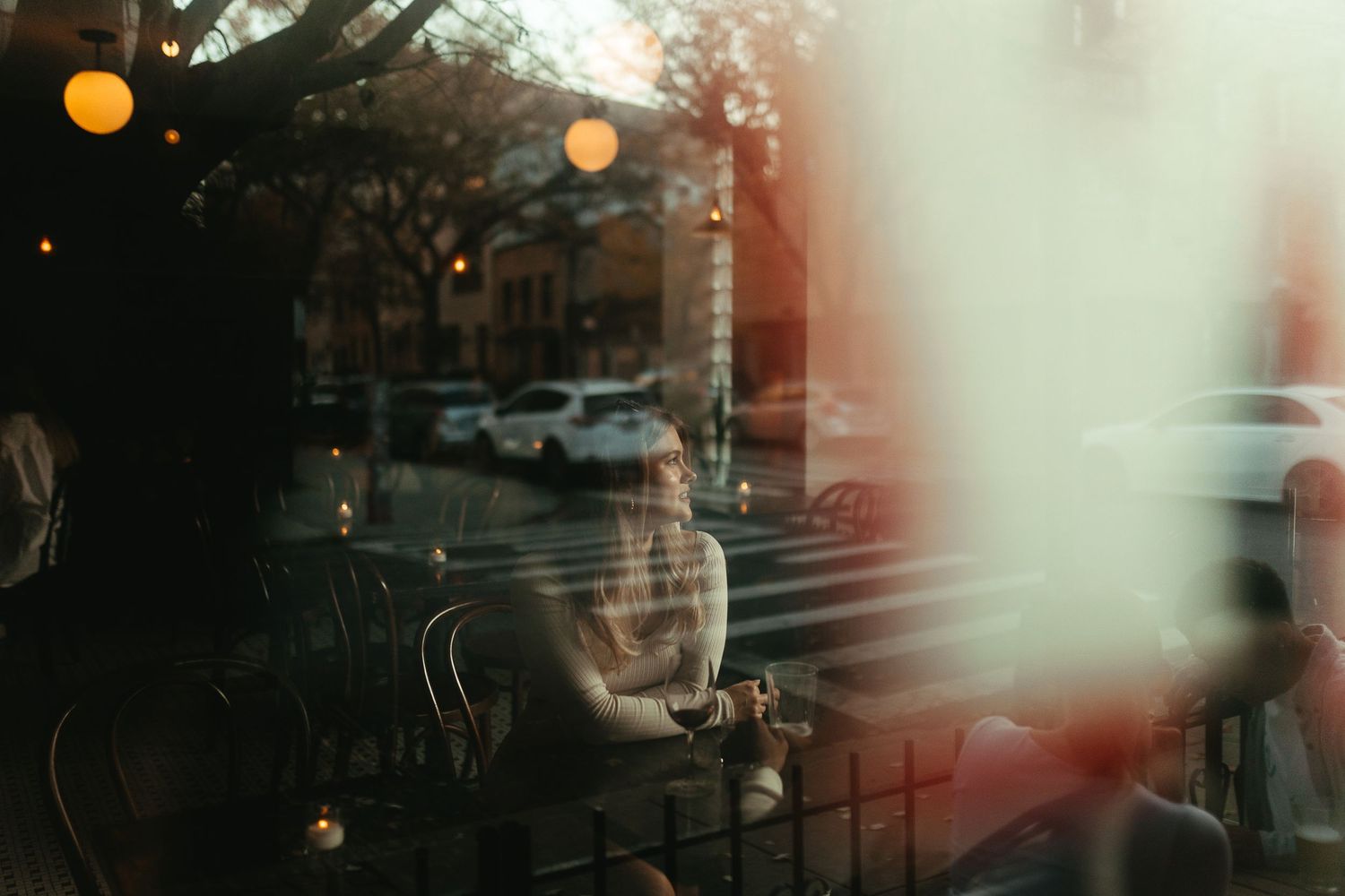 A person sits in a warmly lit cafe, viewed through a reflective window with street traffic visible outside.