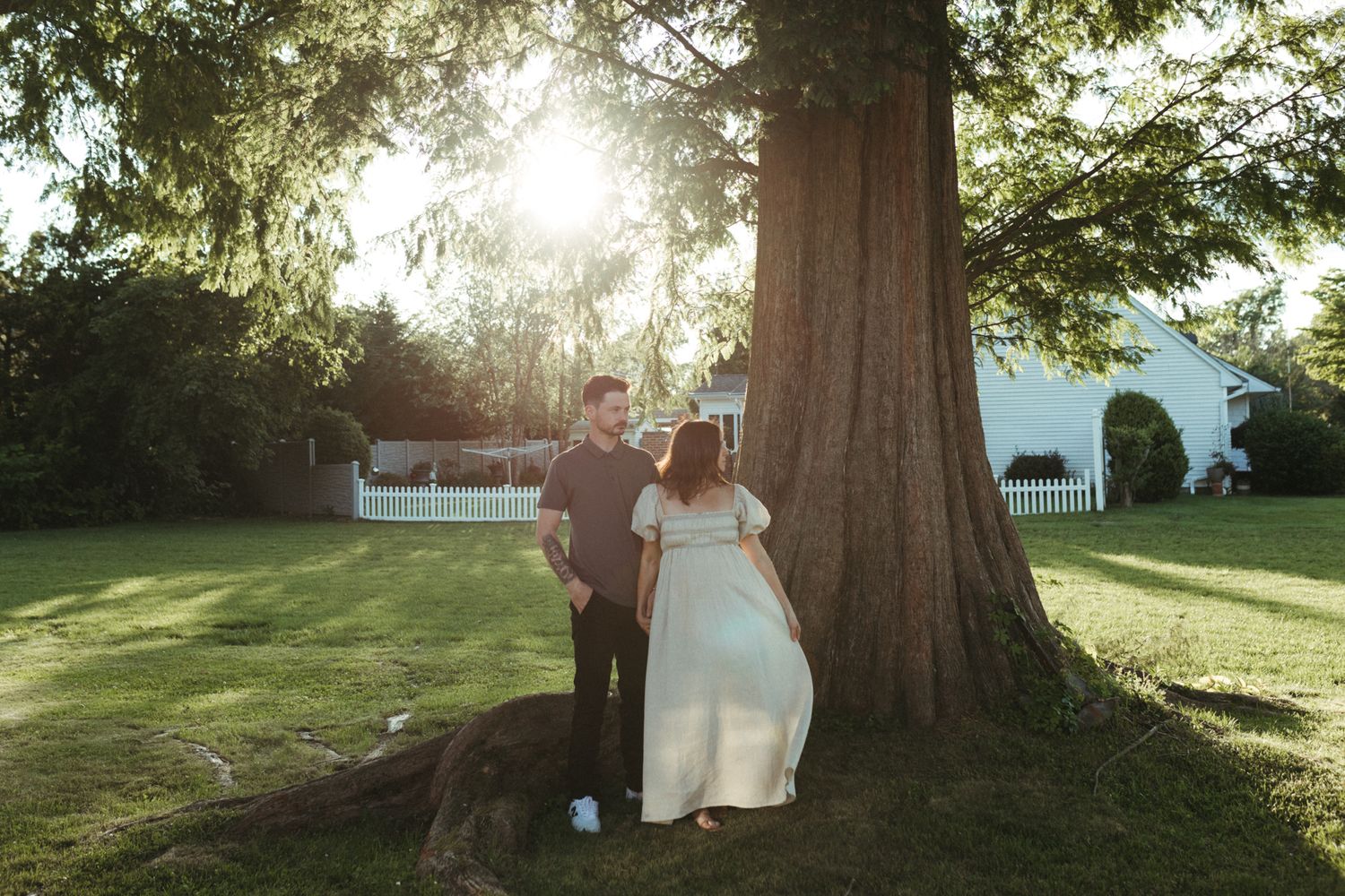 Couple standing near large tree in sunlit backyard, woman in white dress, golden hour light filtering through leaves.