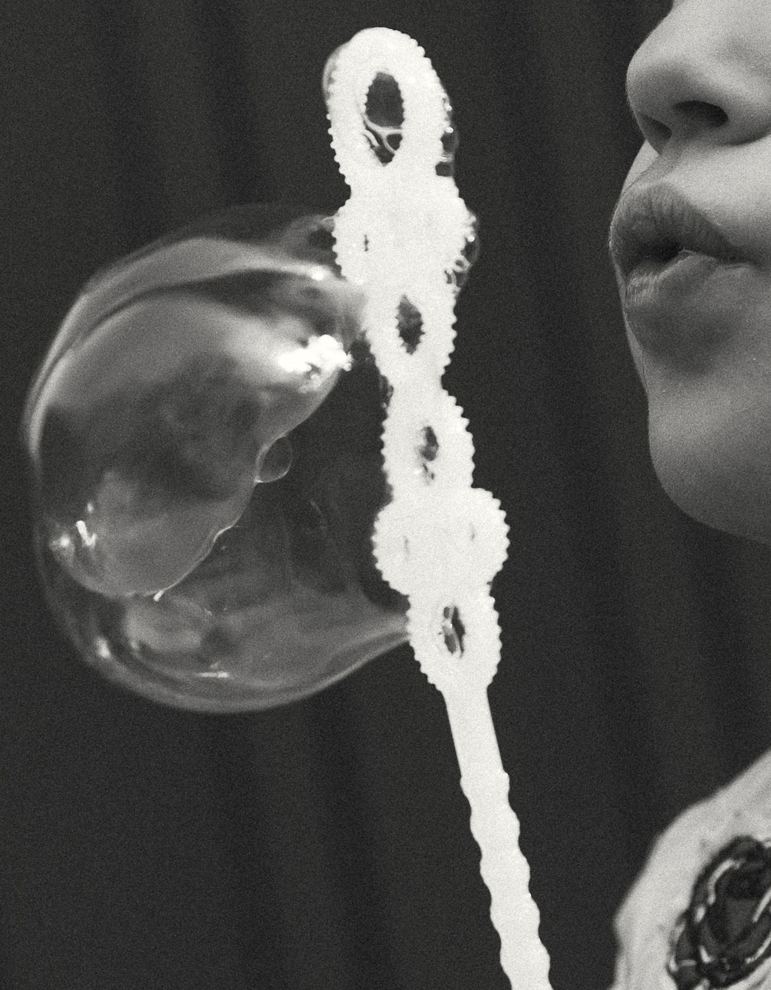 Black and white photo of a child blowing a large soap bubble using a decorative bubble wand.