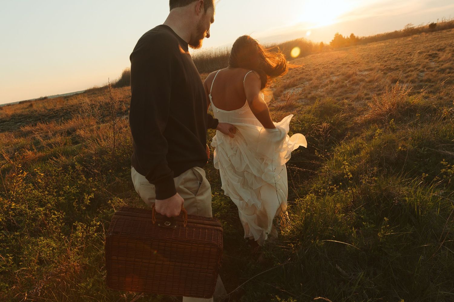 Couple walking through golden field at sunset, man carrying vintage suitcase, woman in flowing white dress.