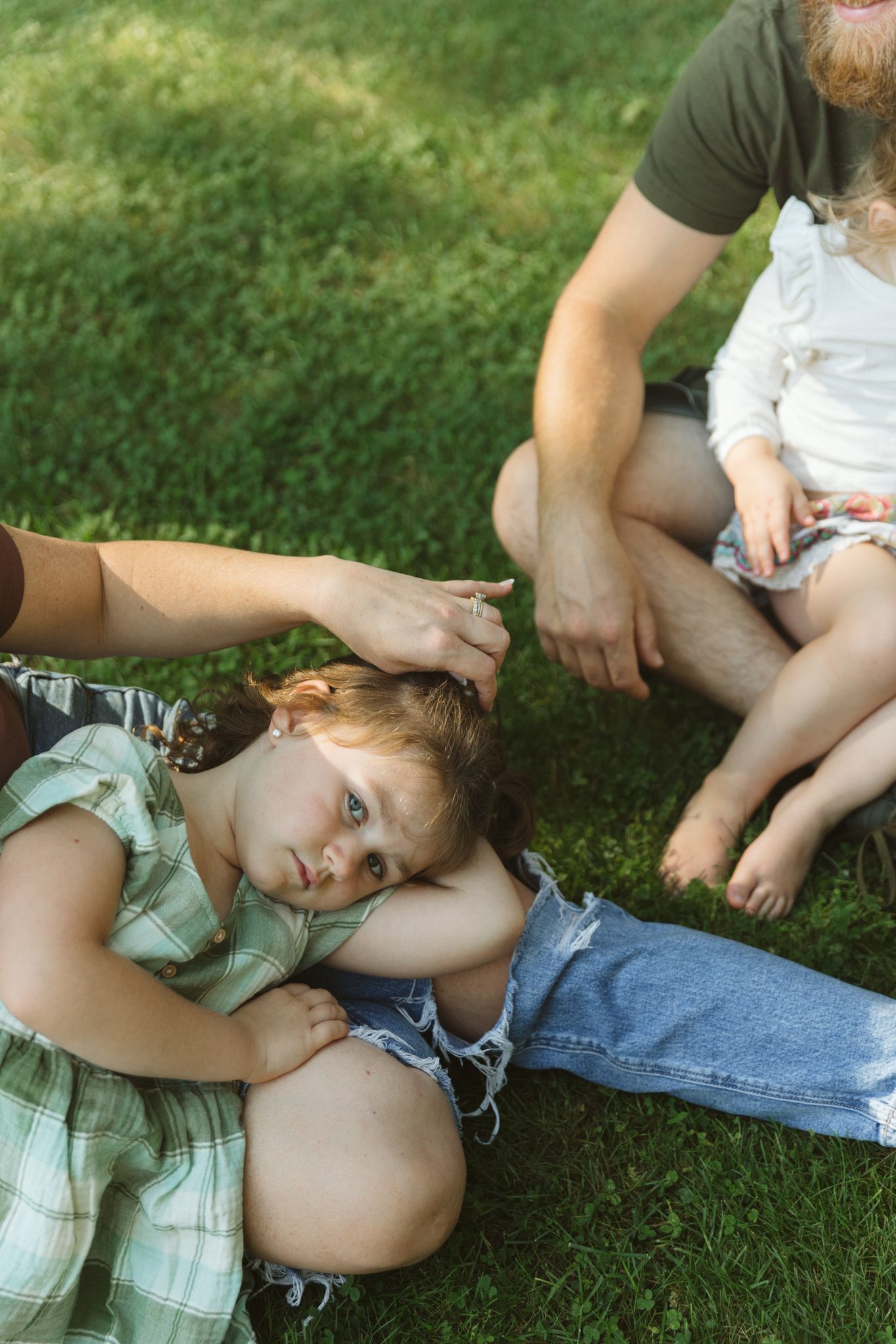 Family relaxing on green grass outdoors, mother braiding young girl's hair while bearded father sits nearby with toddler.
