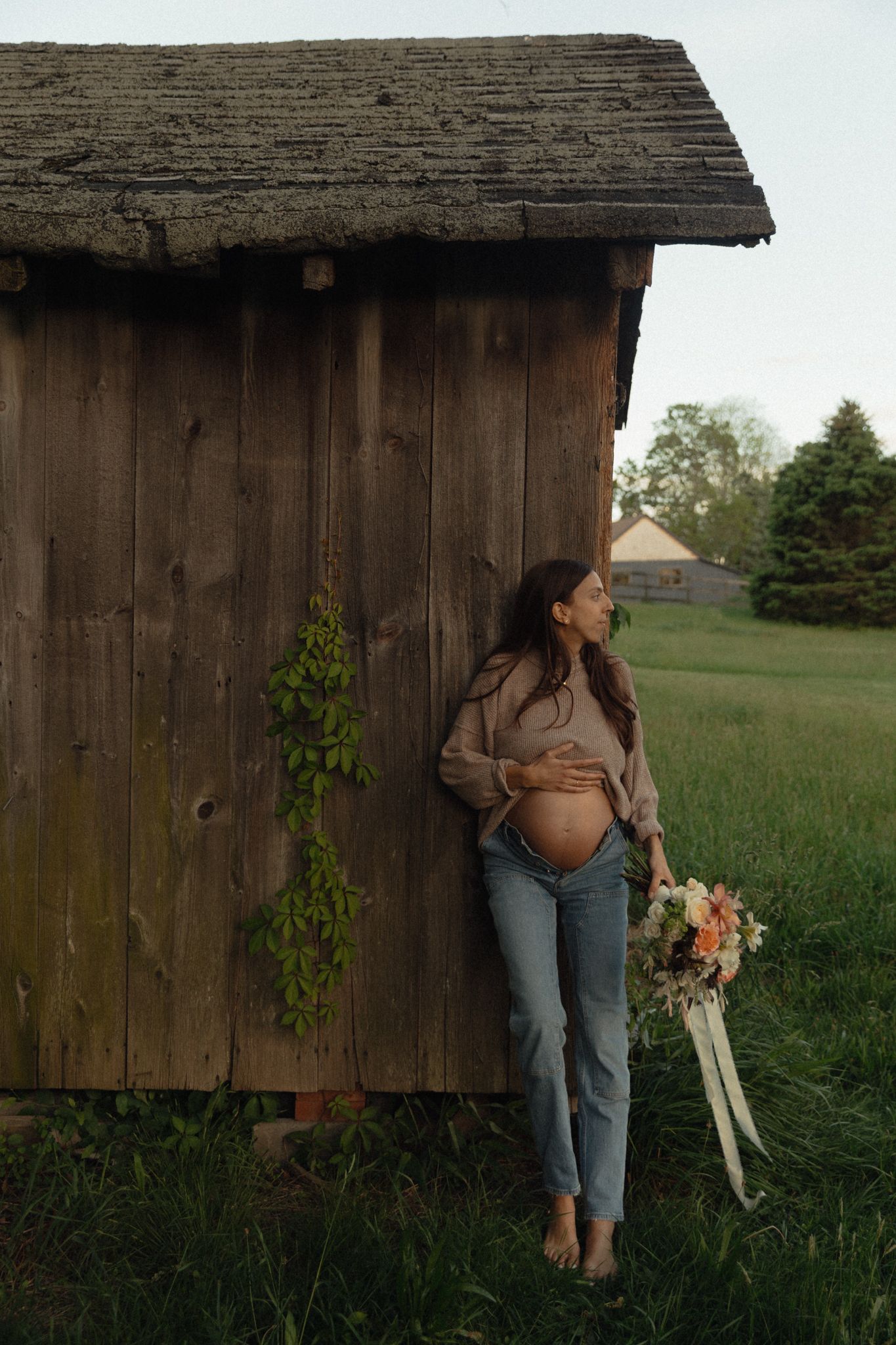 Pregnant woman in blue jeans holds floral bouquet leaning against rustic wooden barn in green countryside.