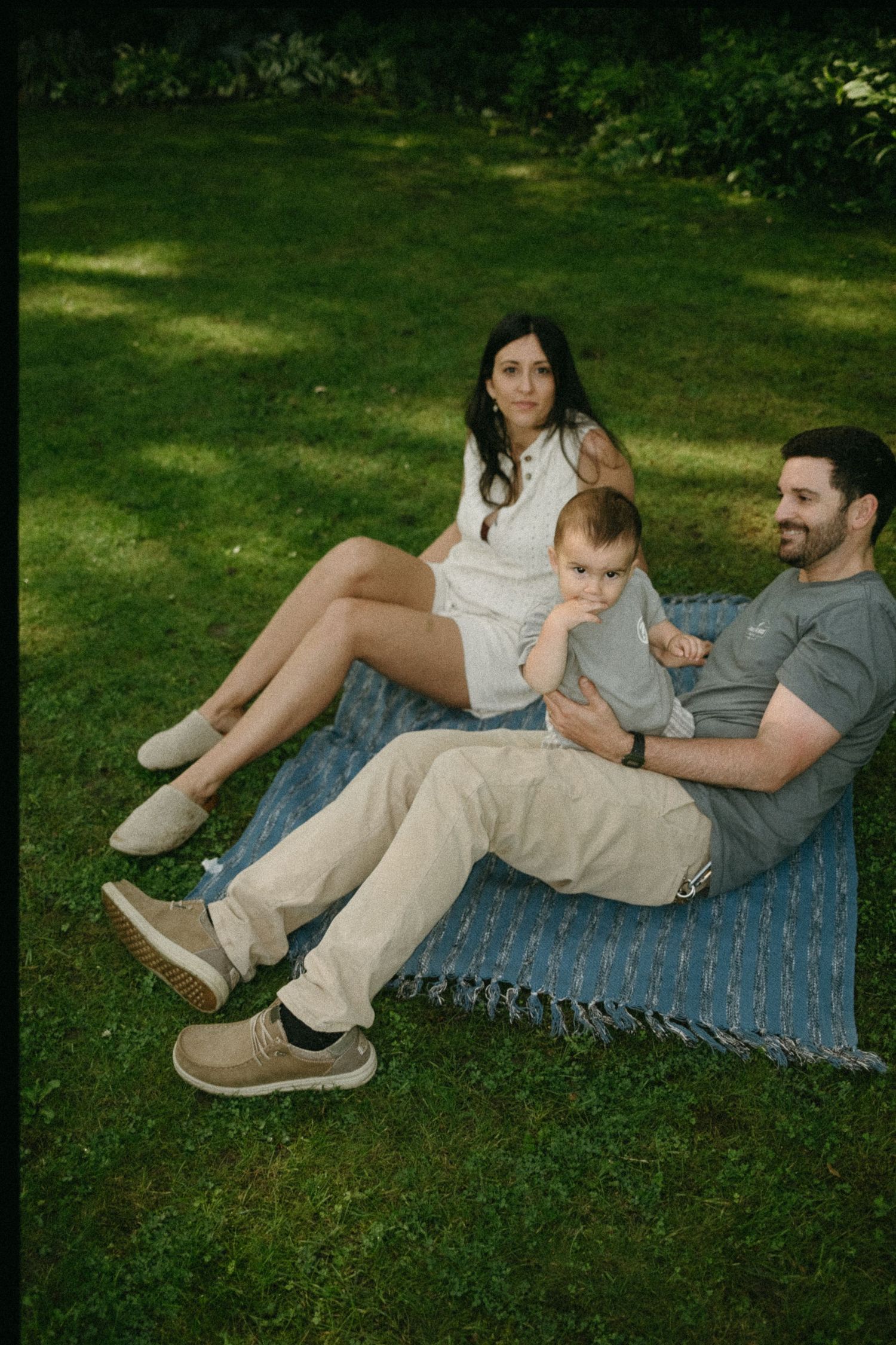 Happy family relaxing on blue blanket in lush green park, parents with baby enjoying outdoor picnic.