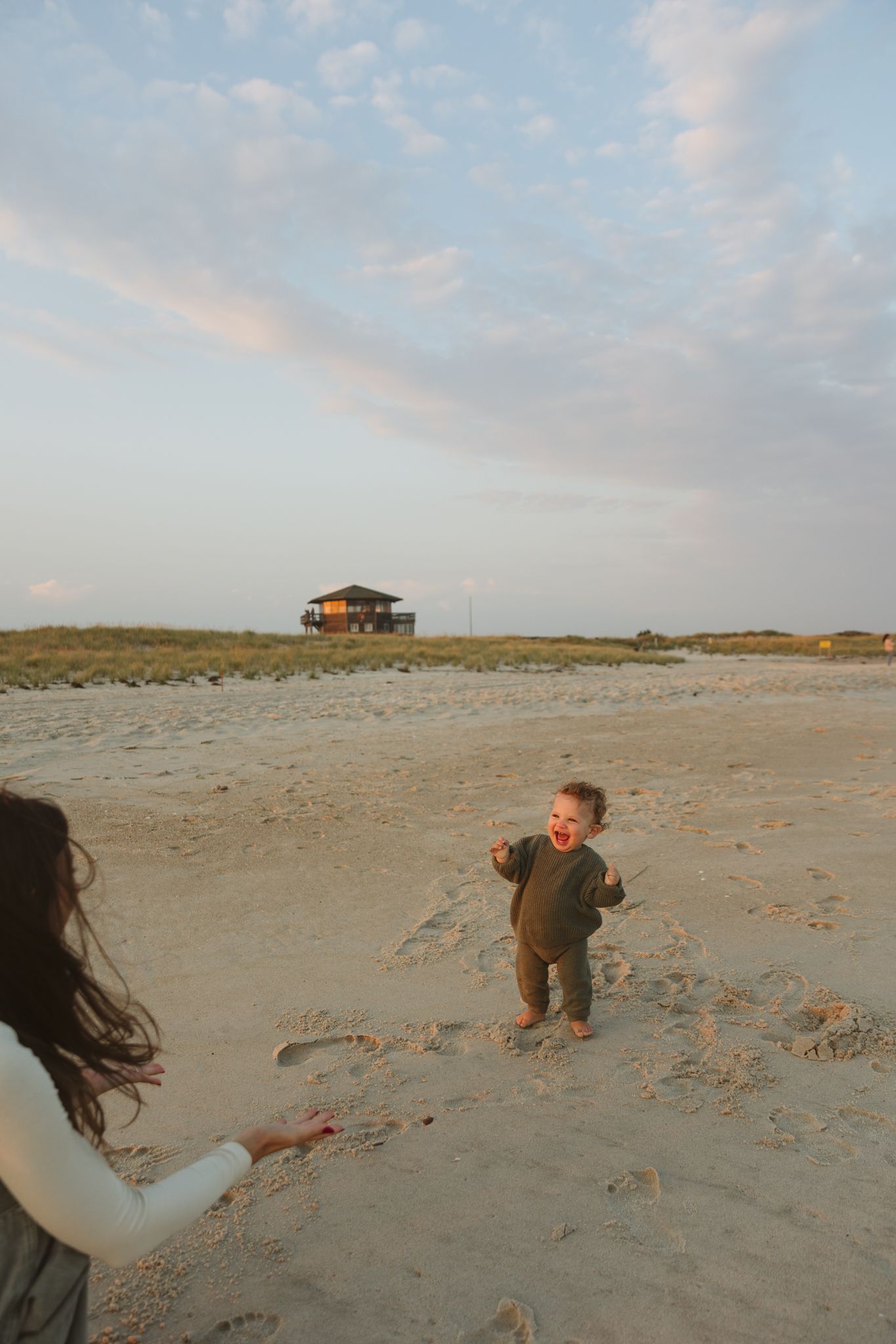 A toddler plays on a beach with a house visible in the distance during sunset.