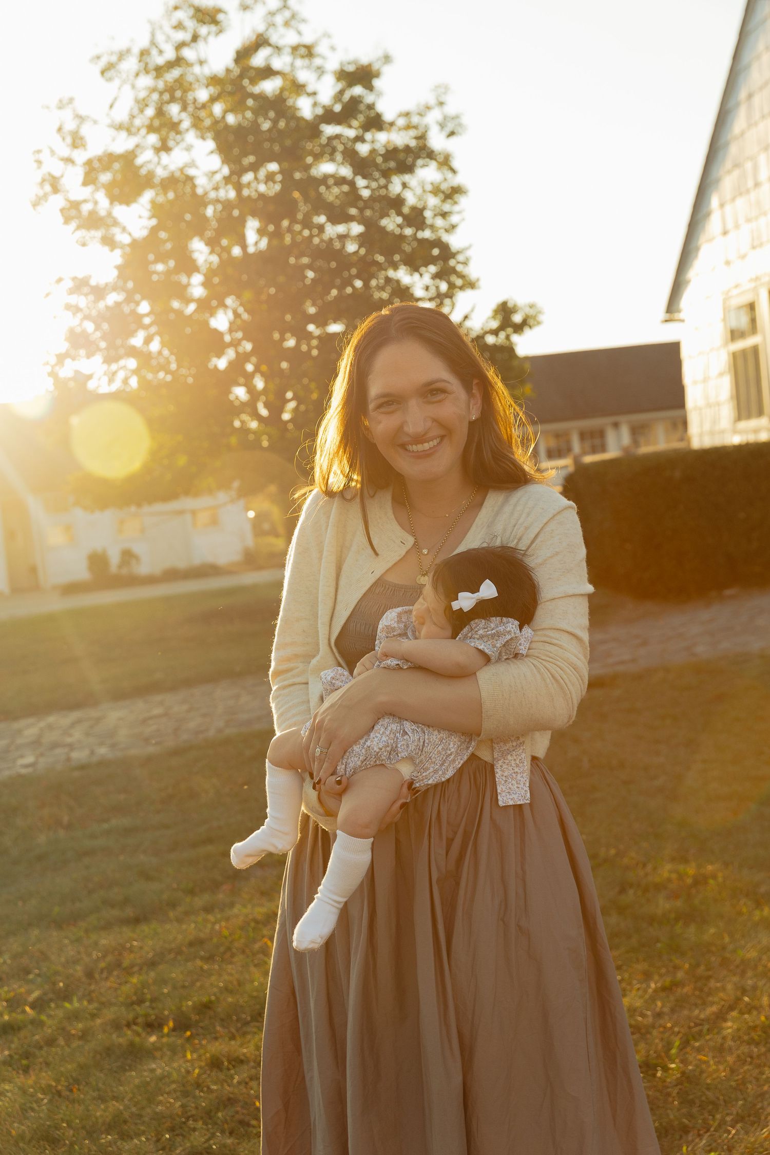 Smiling woman holding a dark-haired baby outdoors at golden hour with warm sunlight and a green tree behind her.