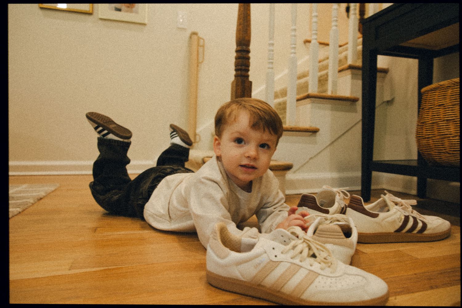 Young toddler lying on hardwood floor next to large white Adidas sneakers near a staircase.