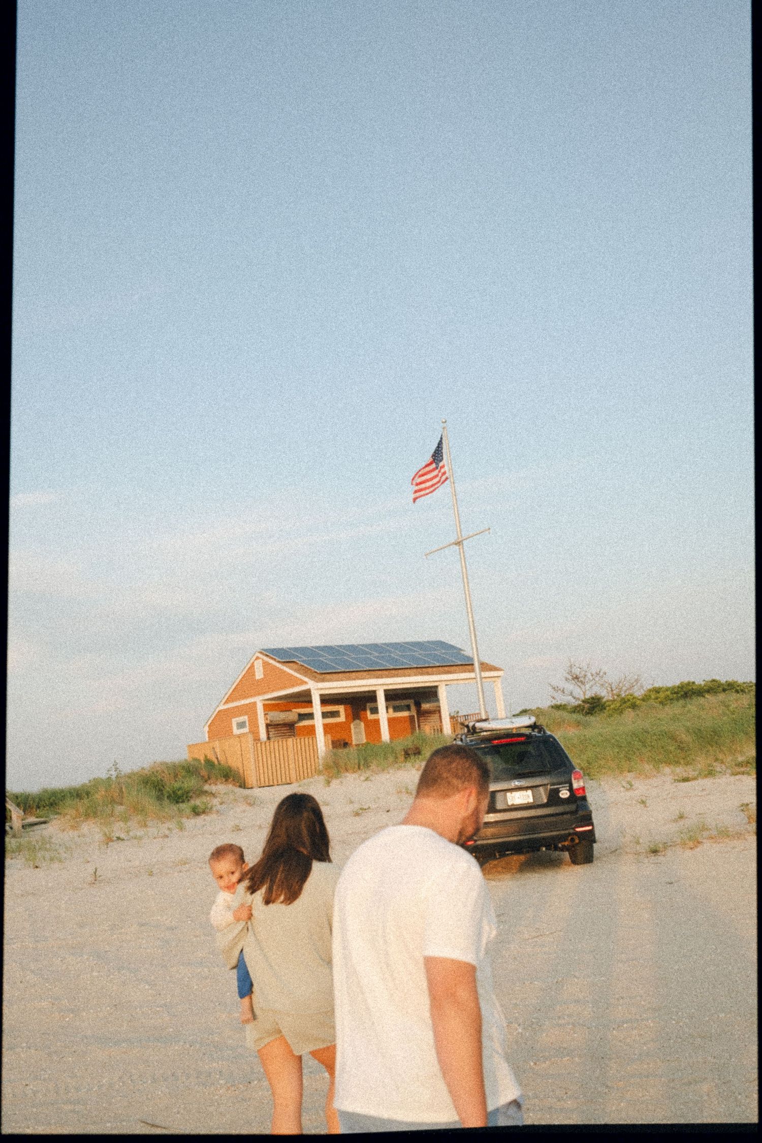 Beach house with American flag and parked car on sandy terrain at dusk.
