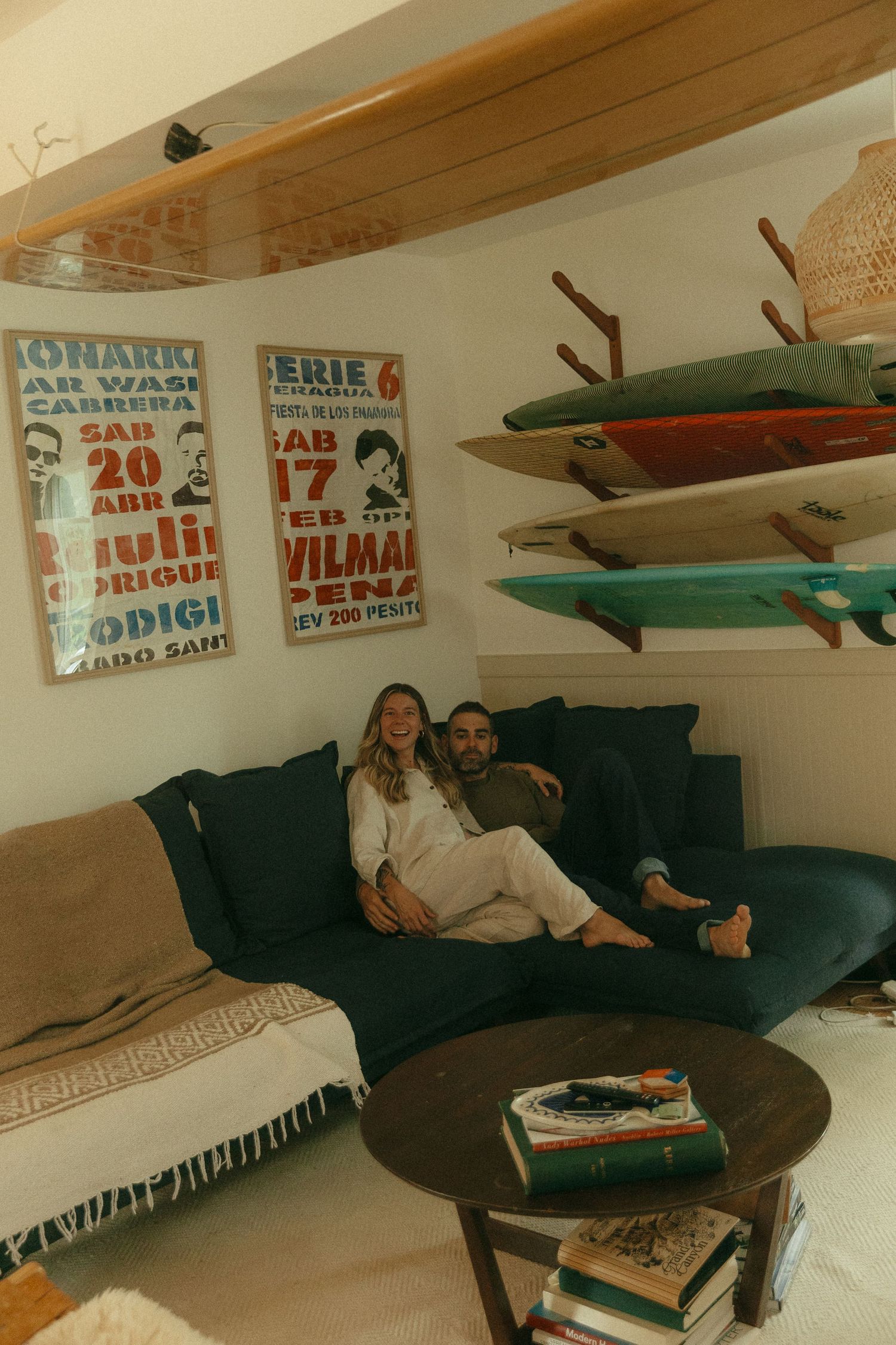 Cozy living room with surfboards mounted on wall, vintage posters, and dark green couch with throw blankets.