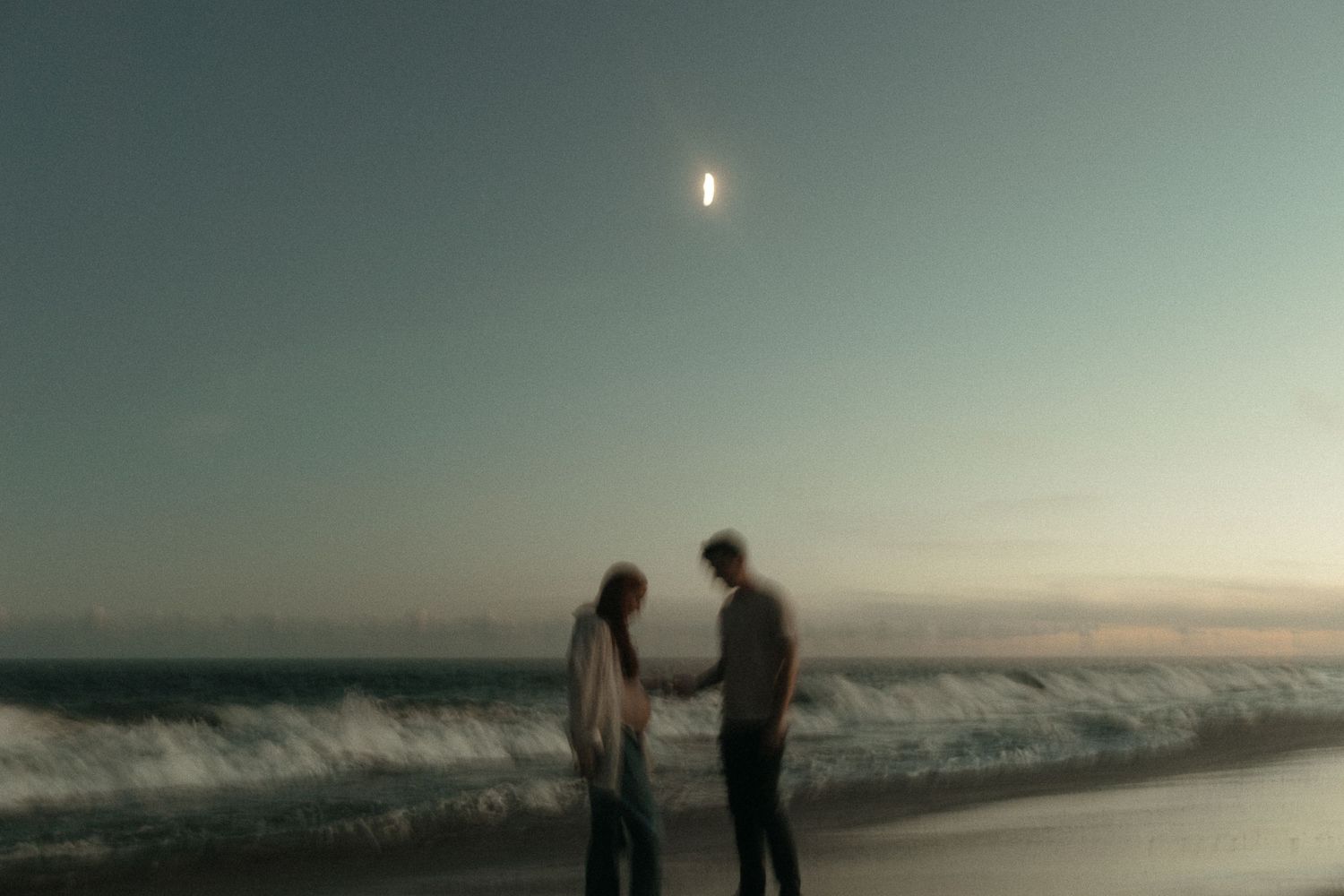 Two figures stand together on a misty beach with the sun glowing in the hazy sky.