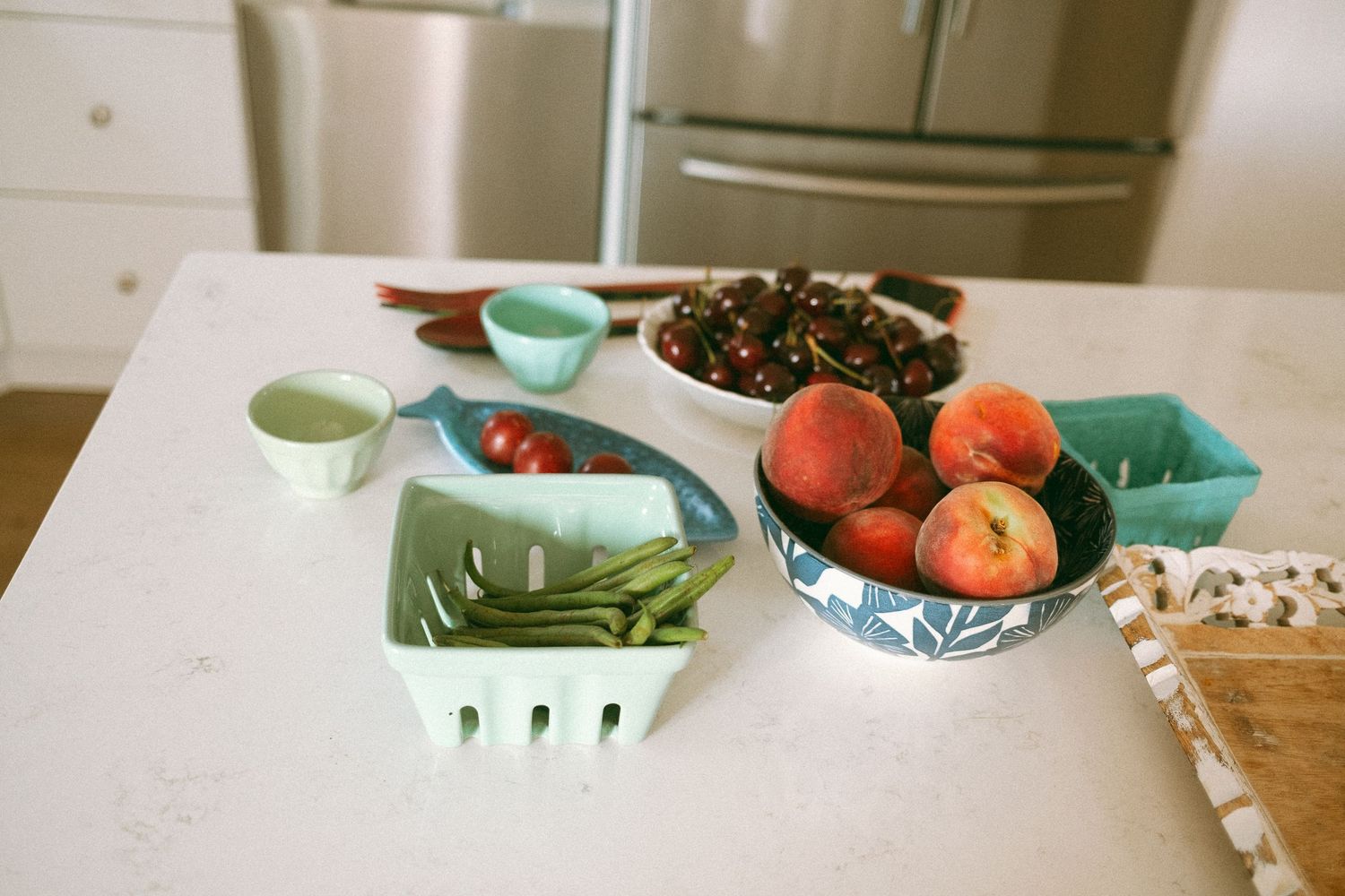Fresh peaches, green beans, tomatoes, and mixed berries arranged in colorful bowls on a white kitchen counter.