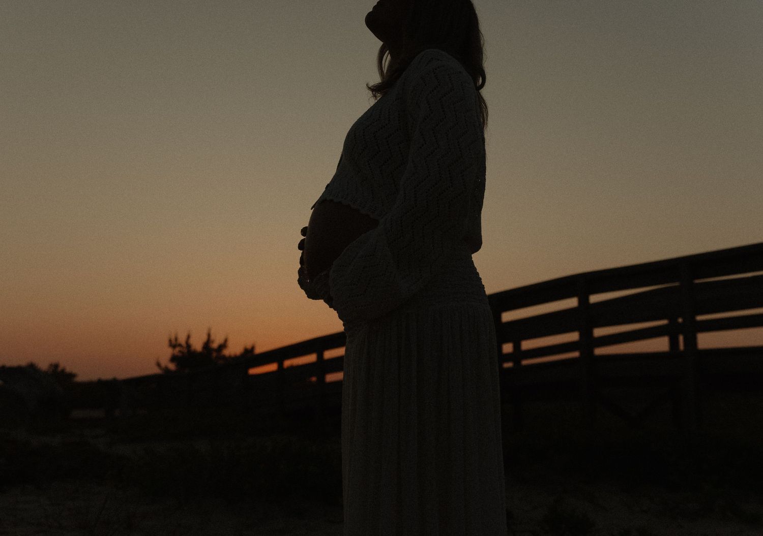 Silhouette of pregnant woman cradling belly at sunset near wooden fence in serene outdoor setting.