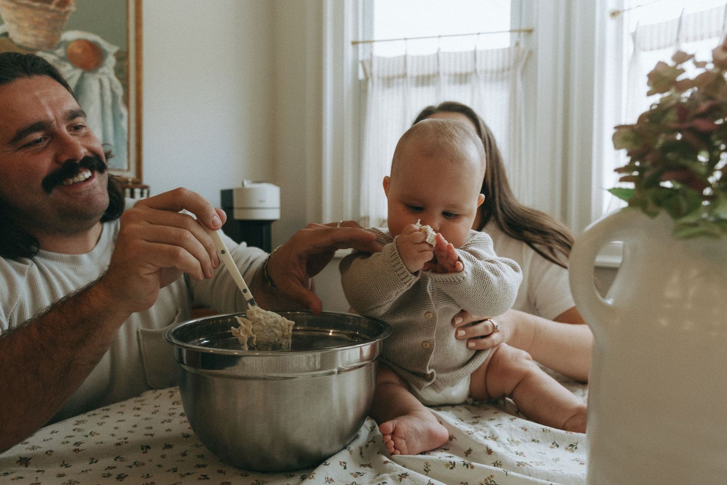 Father and baby mixing ingredients in steel bowl at kitchen table with mother nearby.